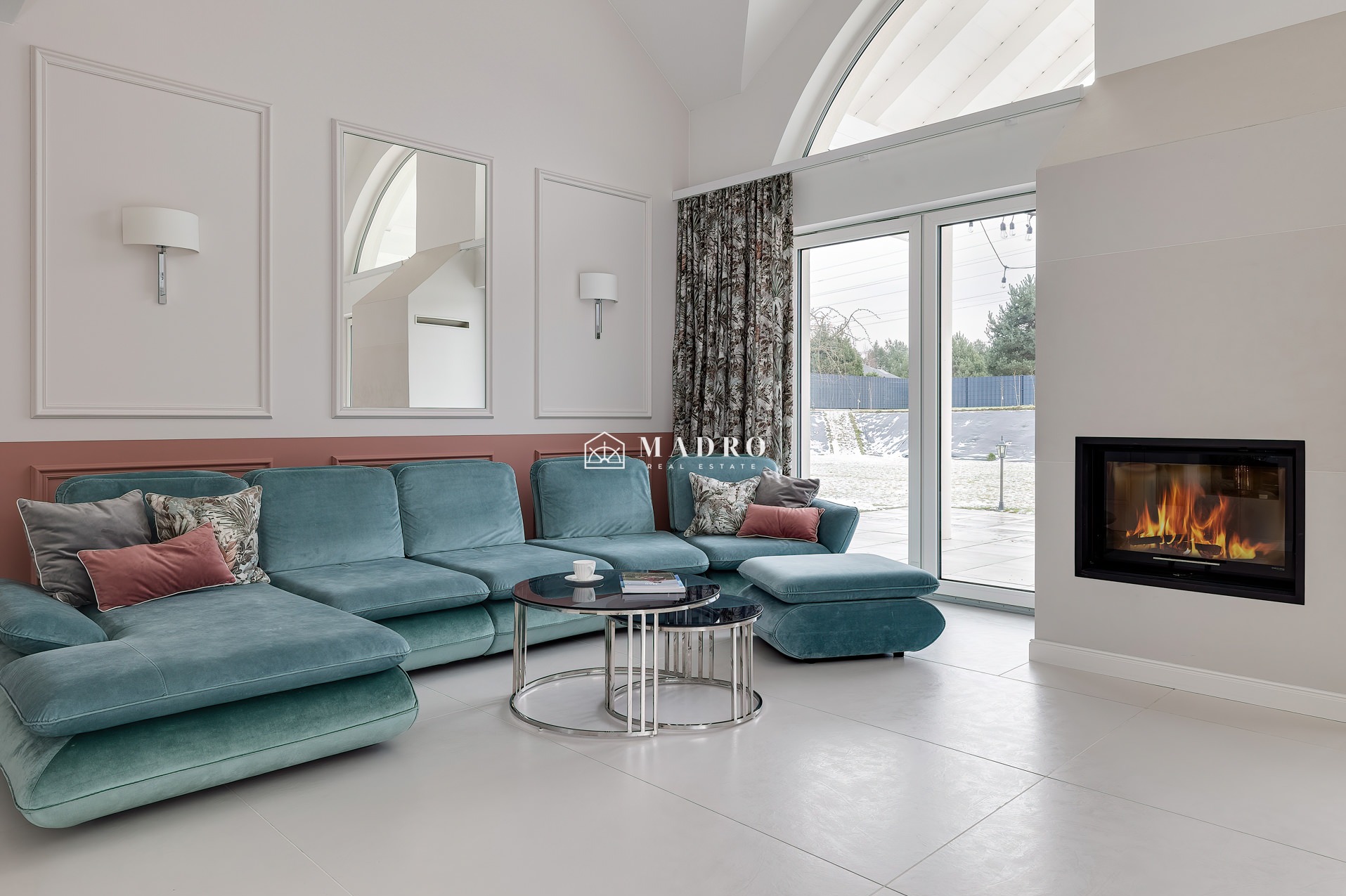 Jasne, przestronne wnętrze salonu z turkusową sofą, okrągłymi stolikami kawowymi, kominkiem z palącym się ogniem i dużym oknem z widokiem na ośnieżony ogród.