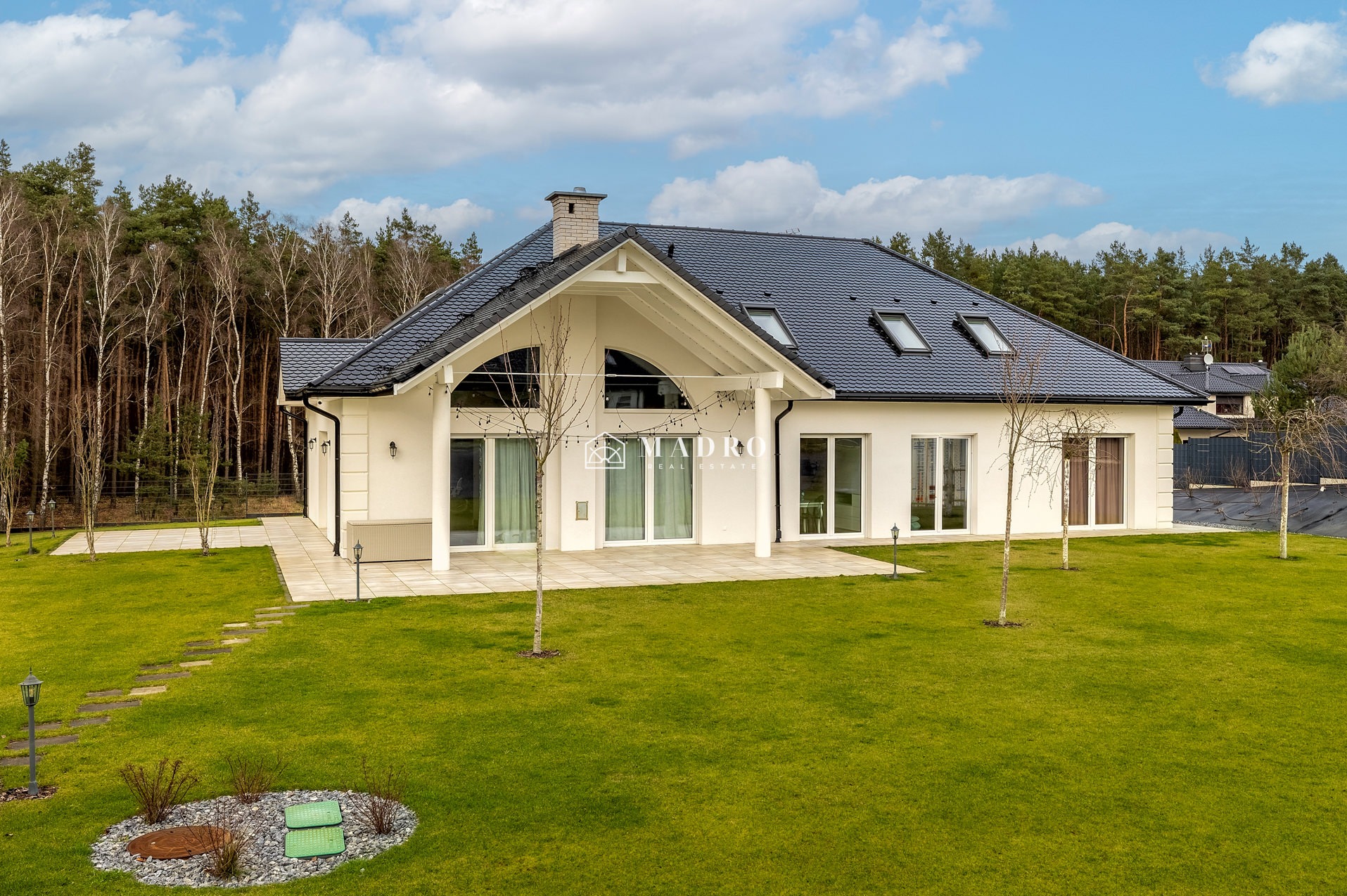 Nowoczesny dom jednorodzinny z białą elewacją, ciemną dachówką i dużym ogrodem, widok z perspektywy ogrodu, na szczycie okno w kształcie półkola.