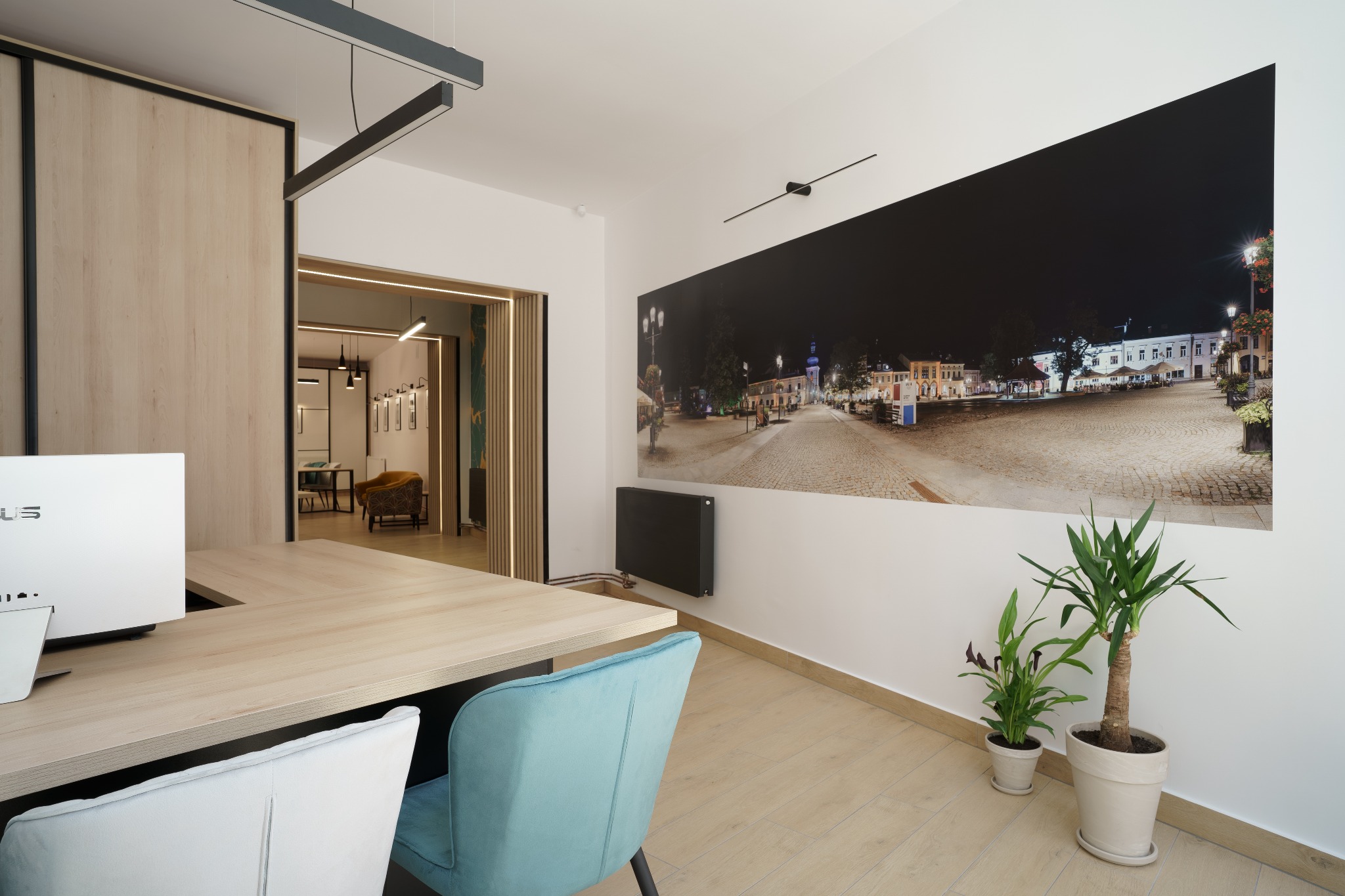 Nowoczesne biuro agencji nieruchomości z biurkiem, krzesłami w kolorze błękitnym i białym, komputerem oraz zdjęciem nocnego rynku miasta na ścianie.