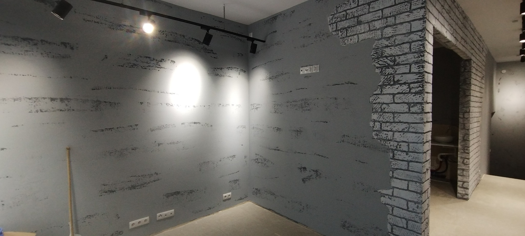 Szara ściana z efektem dekoracyjnym imitującym cegłę przy framudze drzwi, oświetlenie punktowe na suficie.