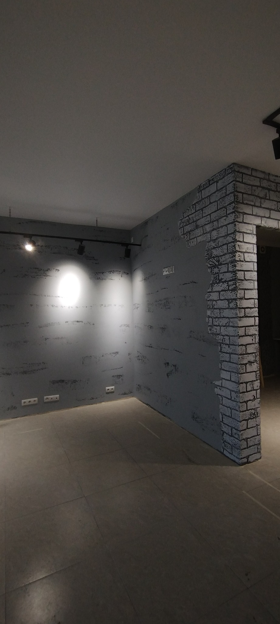 Szare wnętrze z dekoracyjną ścianą imitującą cegłę przy przejściu, ściany wykończone z efektem surowego betonu, widoczne gniazdka elektryczne i oświetlenie sufitowe.