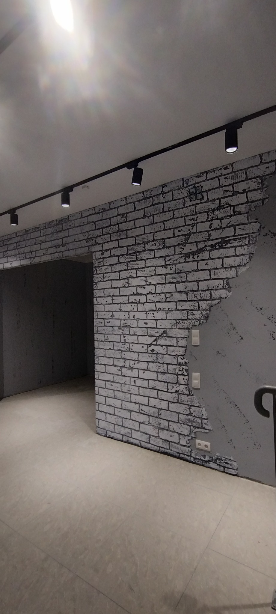Wnętrze z imitacją białej cegły na ścianie, kontrastujące z gładką, szarą powierzchnią i czarnymi reflektorami sufitowymi.