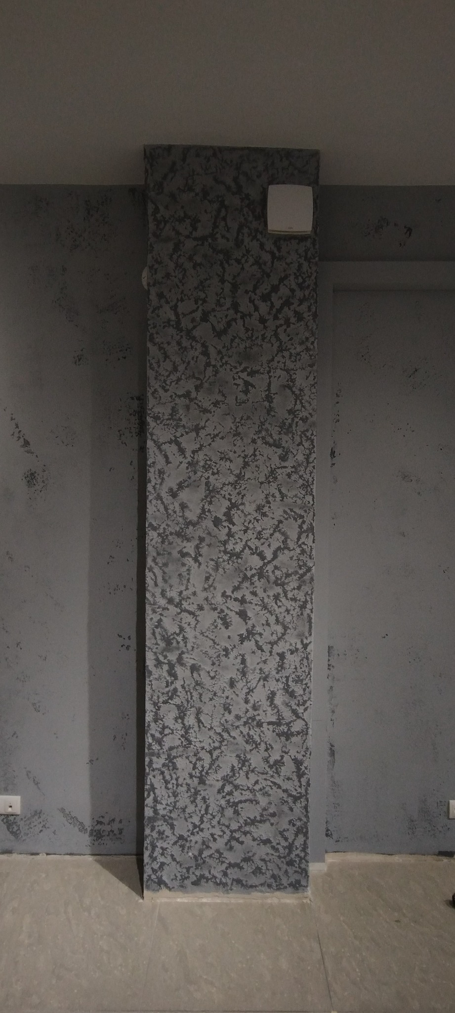 Ściana z dekoracyjną szarą fakturą, obok gładka szara ściana, widoczny fragment podłogi z płytek i biały włącznik na ścianie.
