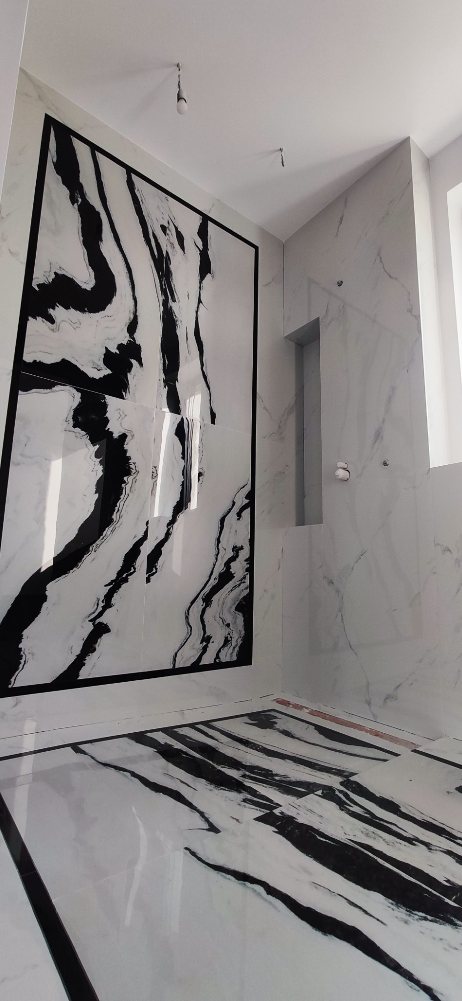 Wykończona łazienka z płytkami imitującymi marmur w kolorach bieli i czerni, wzór żyłkowany na ścianie i podłodze, czarne obramowanie płytek na ścianie, wnęka w ścianie, surowa żarówka na kablu.