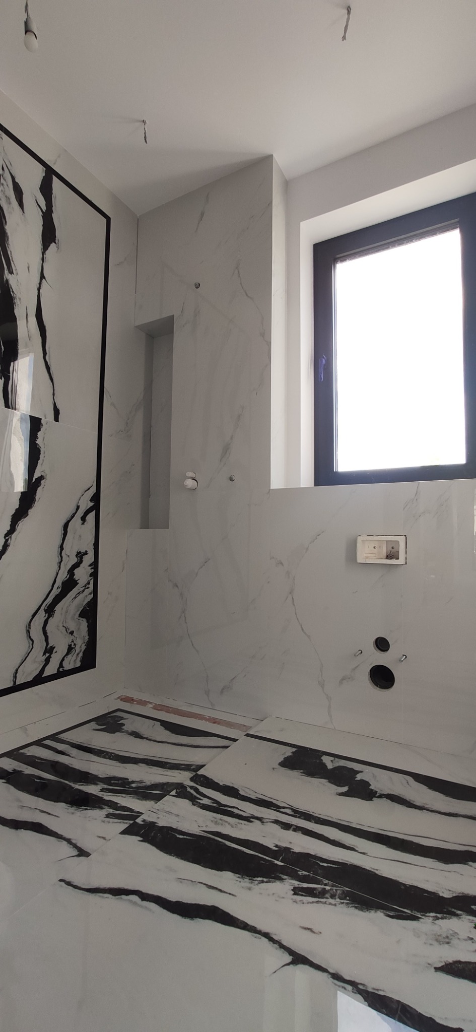 Wnętrze łazienki w trakcie prac wykończeniowych, widoczne płytki imitujące marmur z czarnymi żyłami na ścianach i podłodze, przygotowane otwory na armaturę, okno z czarną ramą, brakujące fugi...