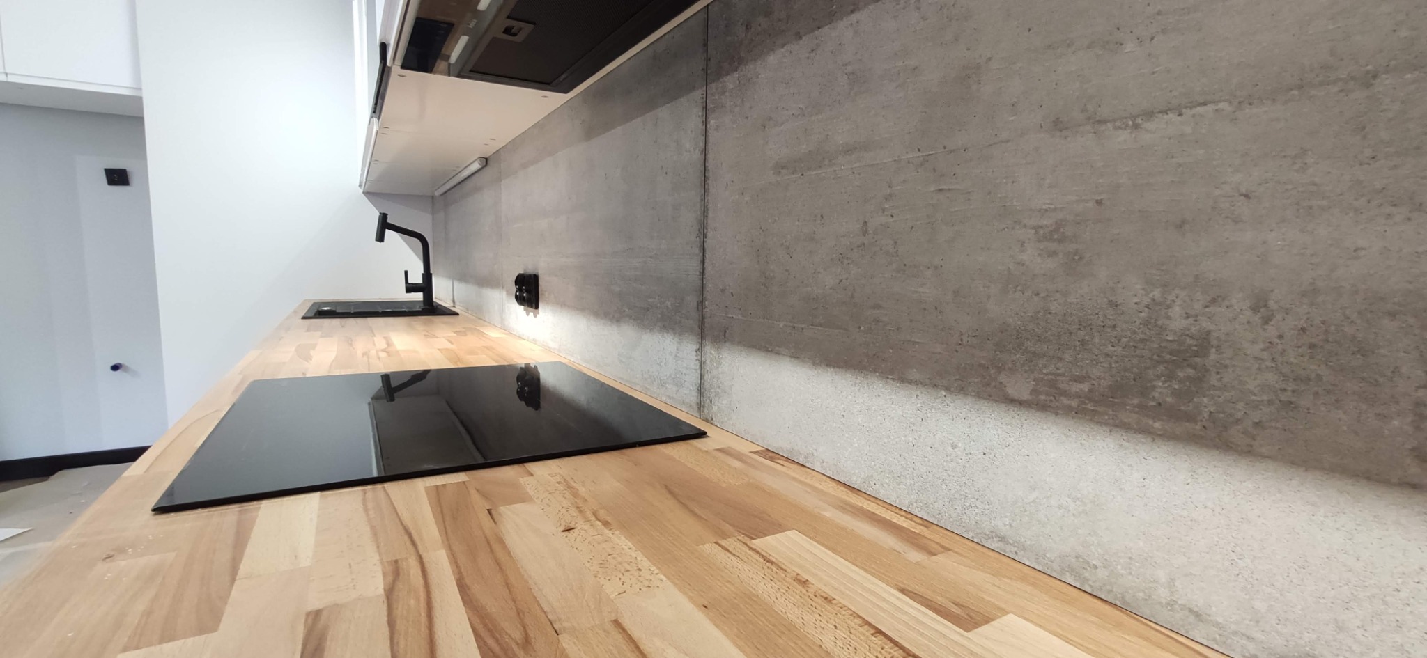 Nowoczesna kuchnia z drewnianym blatem, czarną płytą indukcyjną i baterią oraz betonową ścianą nad blatem.