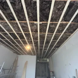 Montaż izolacji z wełny mineralnej w suficie podwieszanym, widoczne drewniane profile konstrukcyjne i surowa ściana w trakcie remontu, wisząca żarówka oświetla przestrzeń.