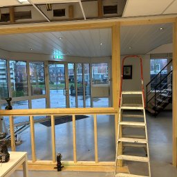 Przemyslaw Krajewski - Montaż drewnianej konstrukcji ścianki działowej w biurze, widoczne narzędzia, poziomica laserowa i drabina oparta o ścianę.