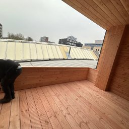 Przemyslaw Krajewski - Pracownik w czarnym ubraniu montuje drewnianą podłogę na tarasie za pomocą wkrętarki akumulatorowej, widok na zabudowę miejską w tle, pochmurne niebo.