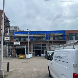 Przemyslaw Krajewski - Budynek komercyjny w trakcie remontu elewacji, osłonięty niebieską folią i rusztowaniami, widoczny z ulicy z zaparkowanymi samochodami i logotypami firm budowlanych.