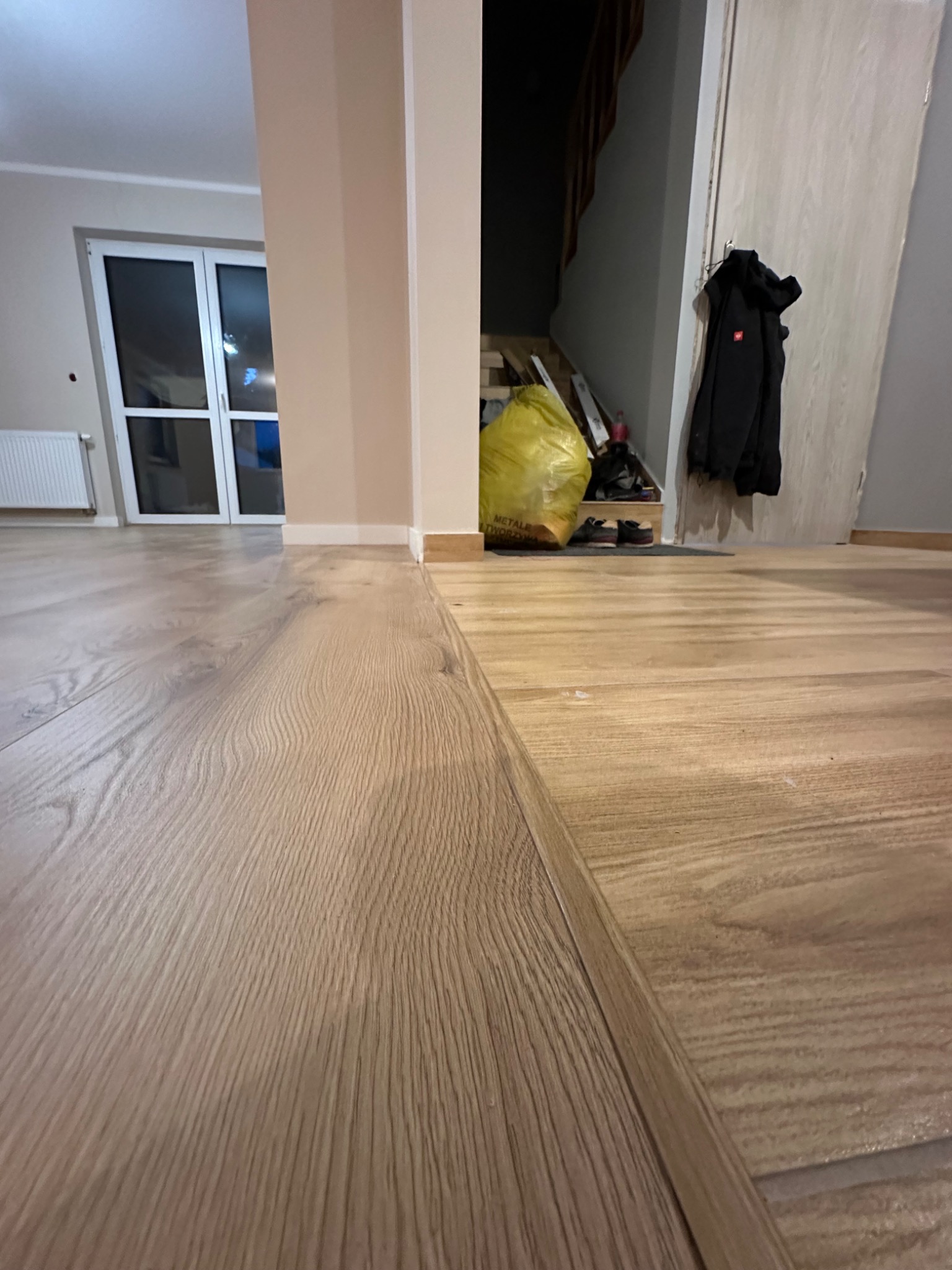 Fragment podłogi z paneli o jasnym odcieniu drewna, widoczne łączenie paneli, w tle drzwi balkonowe, schody i worek z odpadami budowlanymi.