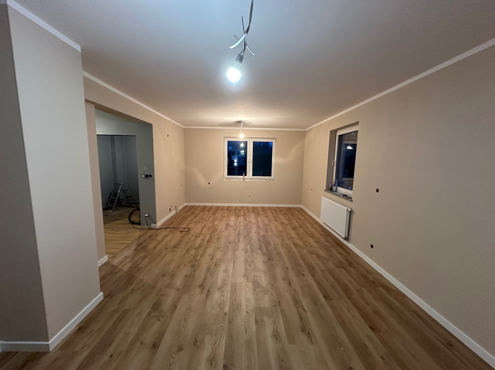 Pusty pokój po remoncie, z panelami podłogowymi imitującymi drewno, świeżo pomalowanymi ścianami w odcieniach beżu i białymi listwami przypodłogowymi. W tle widoczna drabina i kable wystające...