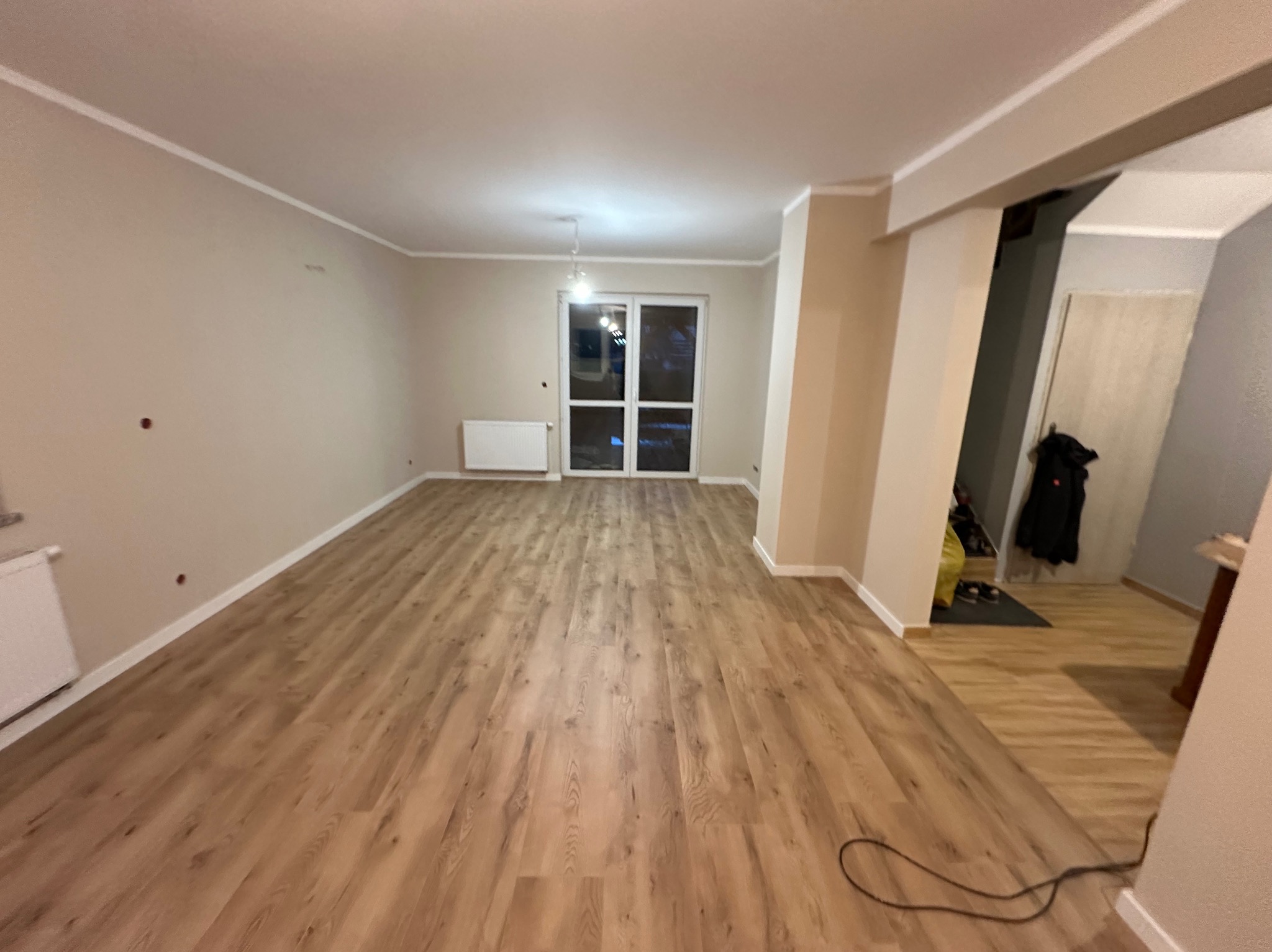 Pusty pokój po remoncie z panelami podłogowymi w odcieniach brązu, widoczne grzejniki, ściany w jasnym beżu z widocznymi śladami po wierceniu, białe listwy przypodłogowe i sufitowe, okno balkonowe...