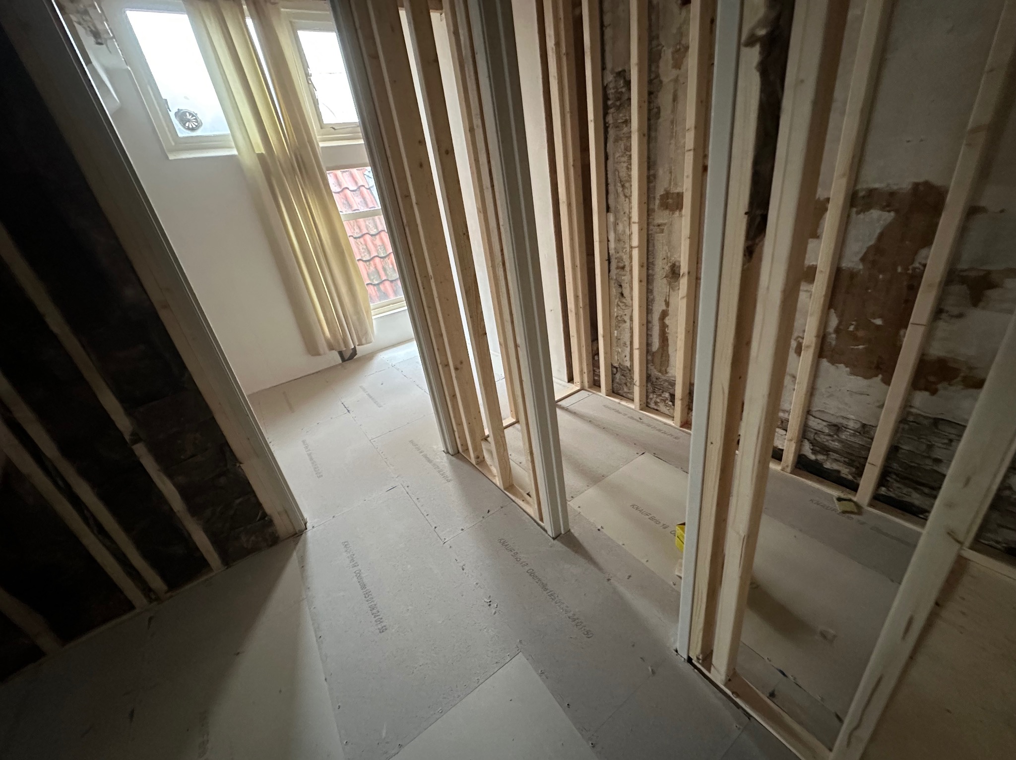 Wnętrze w trakcie prac wykończeniowych, widoczne drewniane stelaże ścian działowych, podłoga pokryta płytami gipsowymi z widocznymi napisami producenta, w tle okno z zasłoną.
