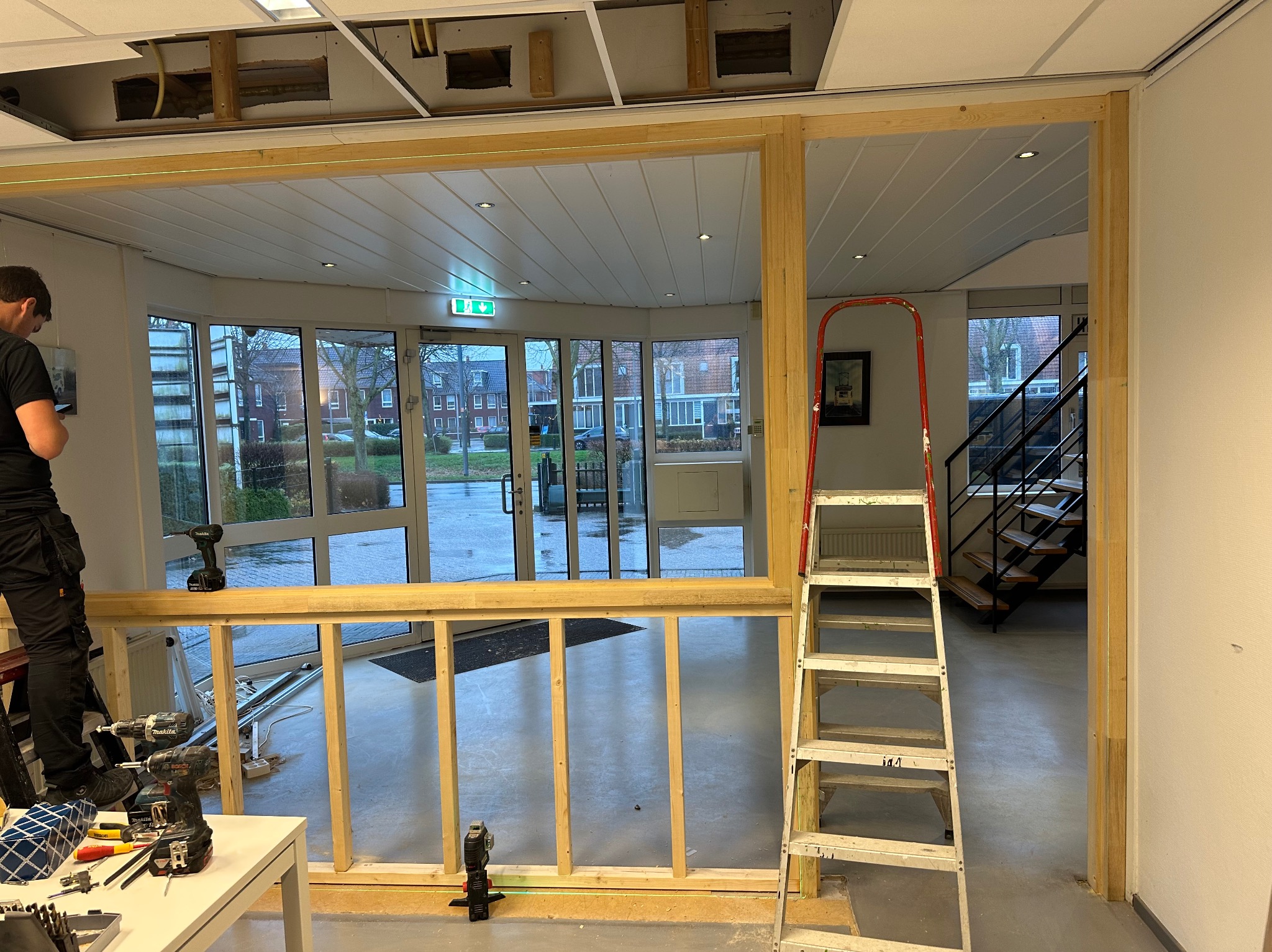 Montaż drewnianej konstrukcji ścianki działowej w biurze, widoczne narzędzia, poziomica laserowa i drabina oparta o ścianę.