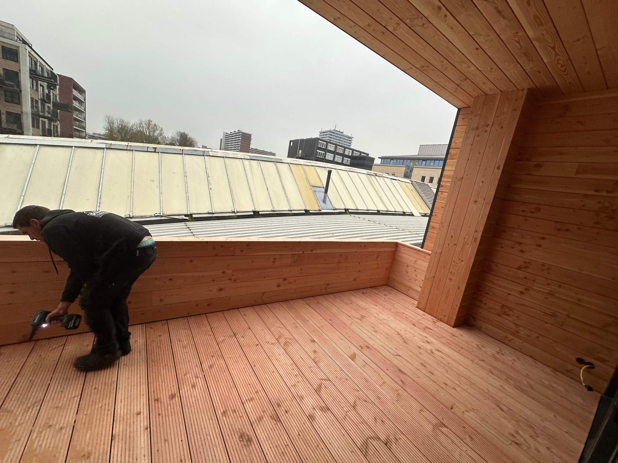 Pracownik w czarnym ubraniu montuje drewnianą podłogę na tarasie za pomocą wkrętarki akumulatorowej, widok na zabudowę miejską w tle, pochmurne niebo.