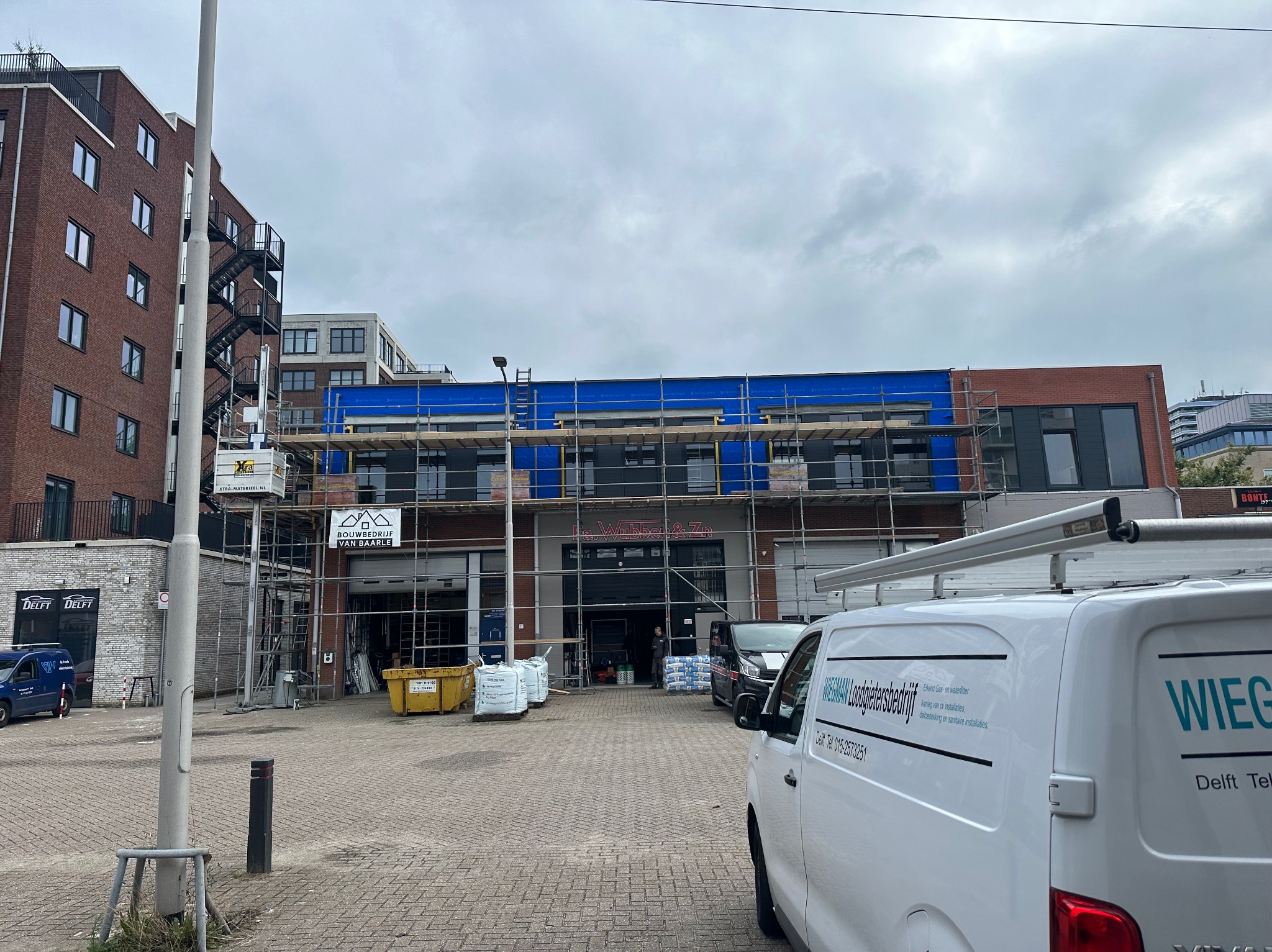 Budynek komercyjny w trakcie remontu elewacji, osłonięty niebieską folią i rusztowaniami, widoczny z ulicy z zaparkowanymi samochodami i logotypami firm budowlanych.