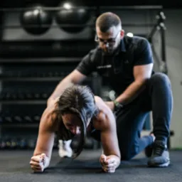 Kobieta w pozycji plank, ćwicząca pod nadzorem mężczyzny w siłowni w Gdańsku. Trener koryguje postawę, dbając o prawidłowe ułożenie pleców.