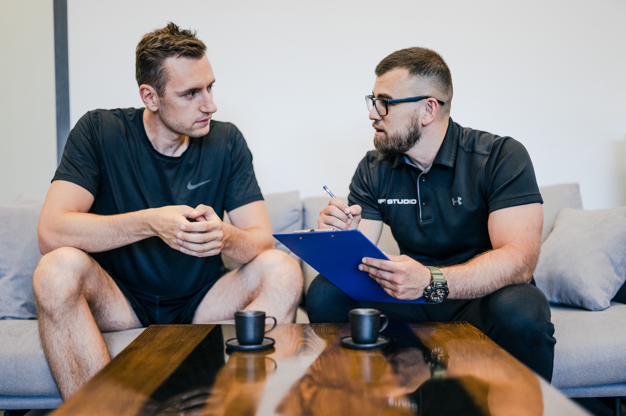 Mężczyzna w sportowym stroju siedzi na kanapie i rozmawia z mężczyzną w okularach, który trzyma niebieski clipboard i długopis; na stoliku kawowym stoją dwie czarne filiżanki.