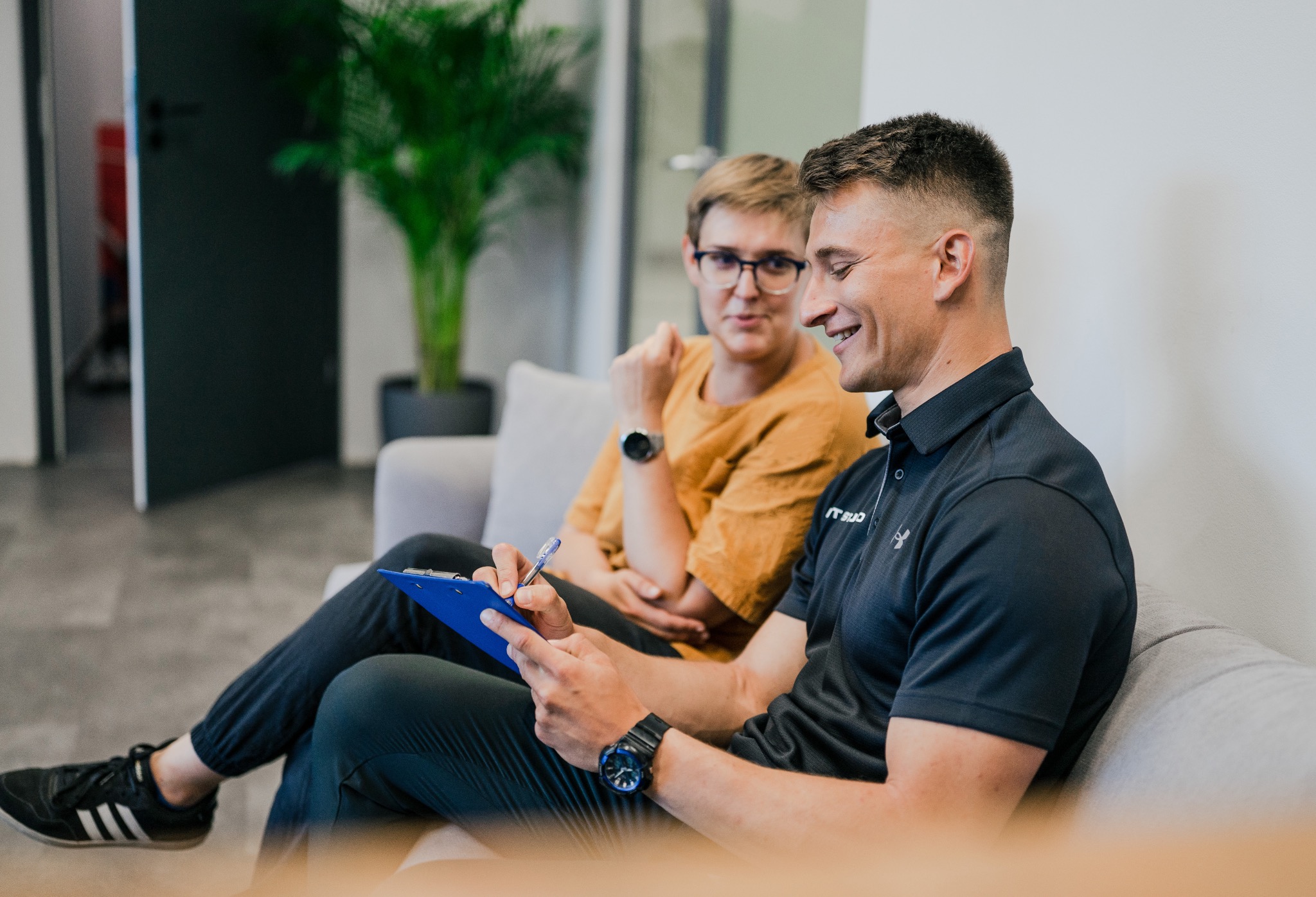 Mężczyzna w czarnej koszulce polo z logo, siedzący na szarej kanapie i wypełniający niebieski formularz na podkładce, uśmiecha się do klienta w okularach siedzącego obok.
