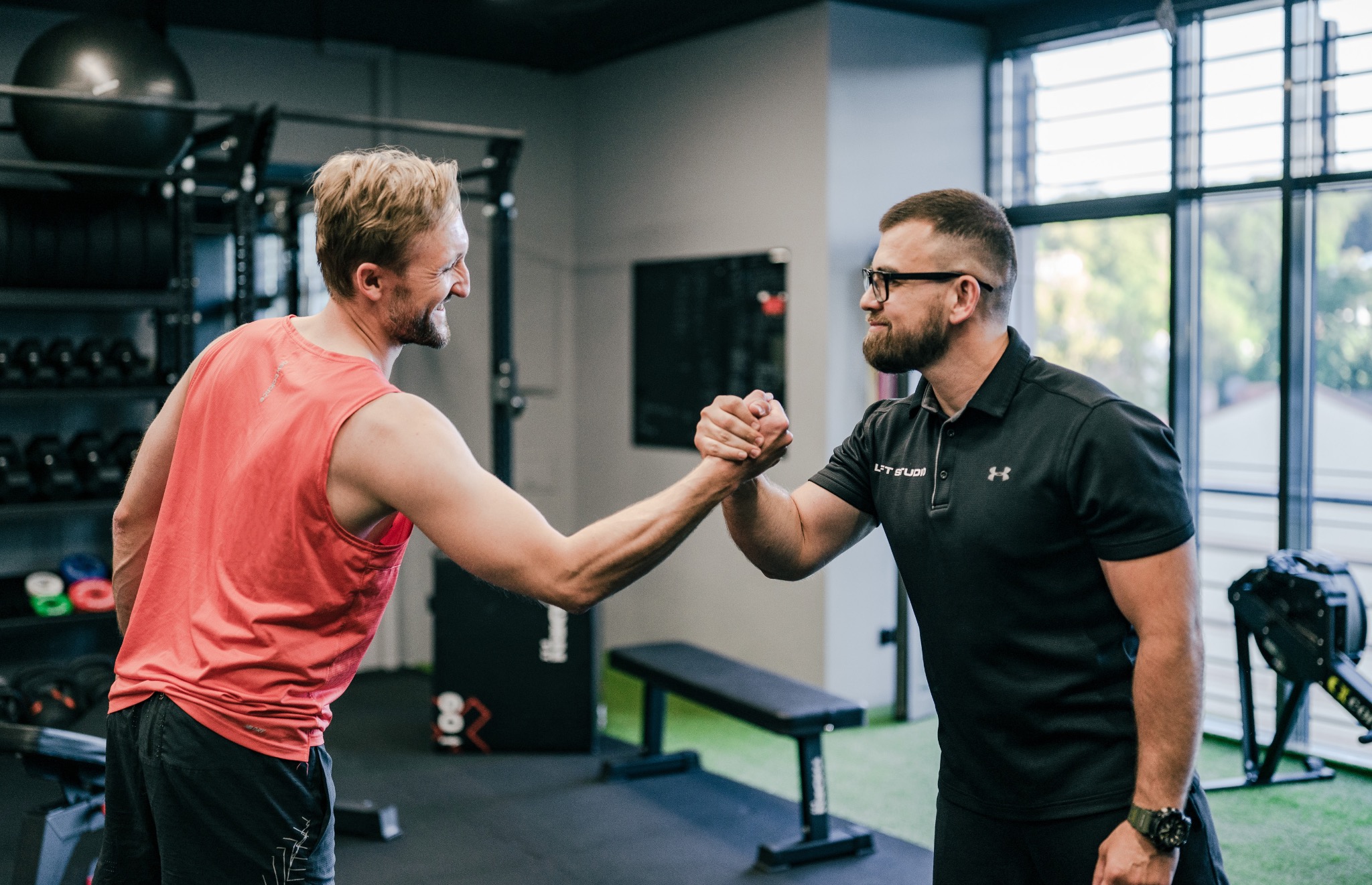 Dwóch mężczyzn w sportowych strojach ściska sobie dłonie w siłowni, z uśmiechami na twarzach, sprzęt do ćwiczeń i duże okno w tle.