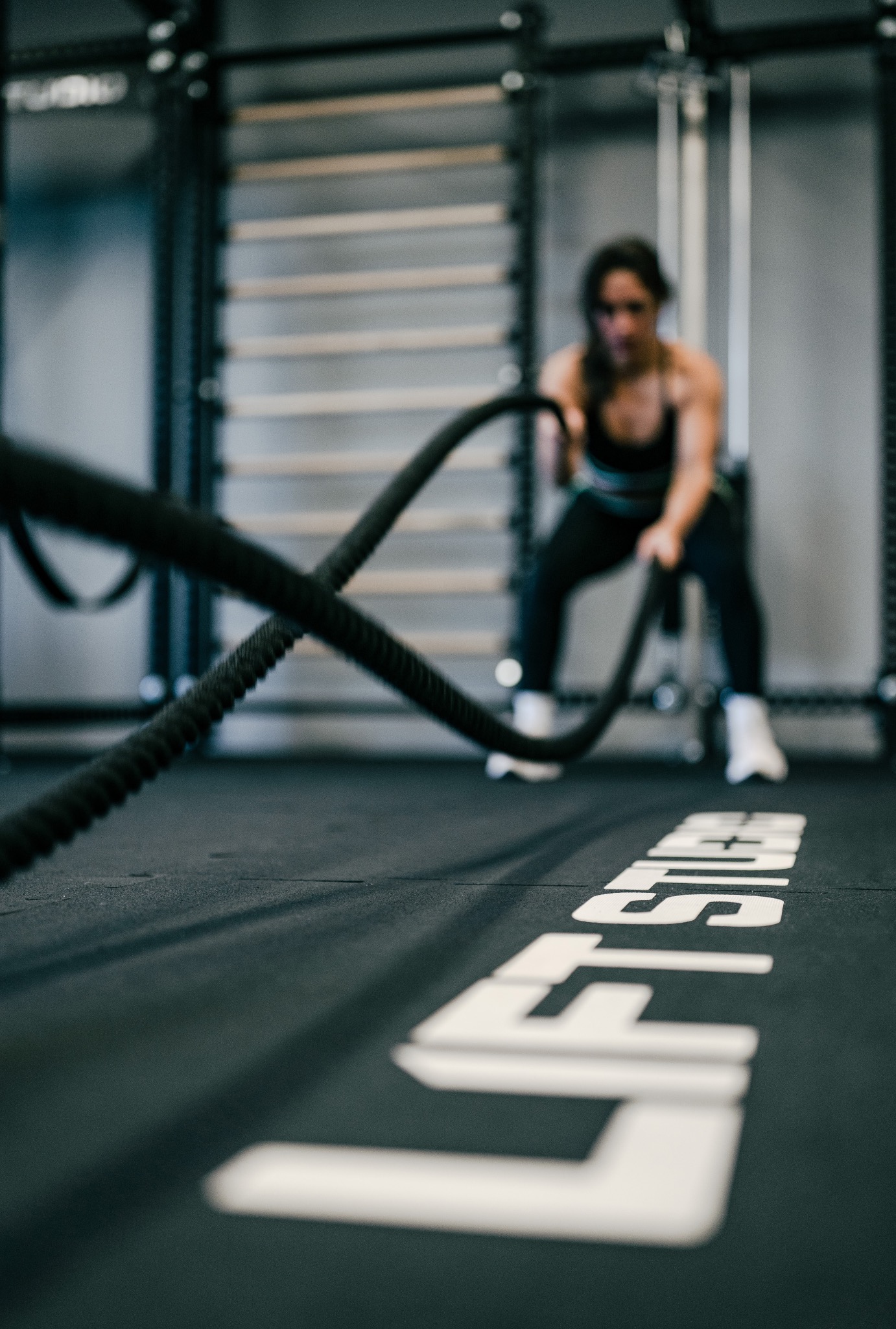 Kobieta ćwiczy z linami treningowymi w siłowni, widoczne logo na podłodze, drabinki gimnastyczne w tle.