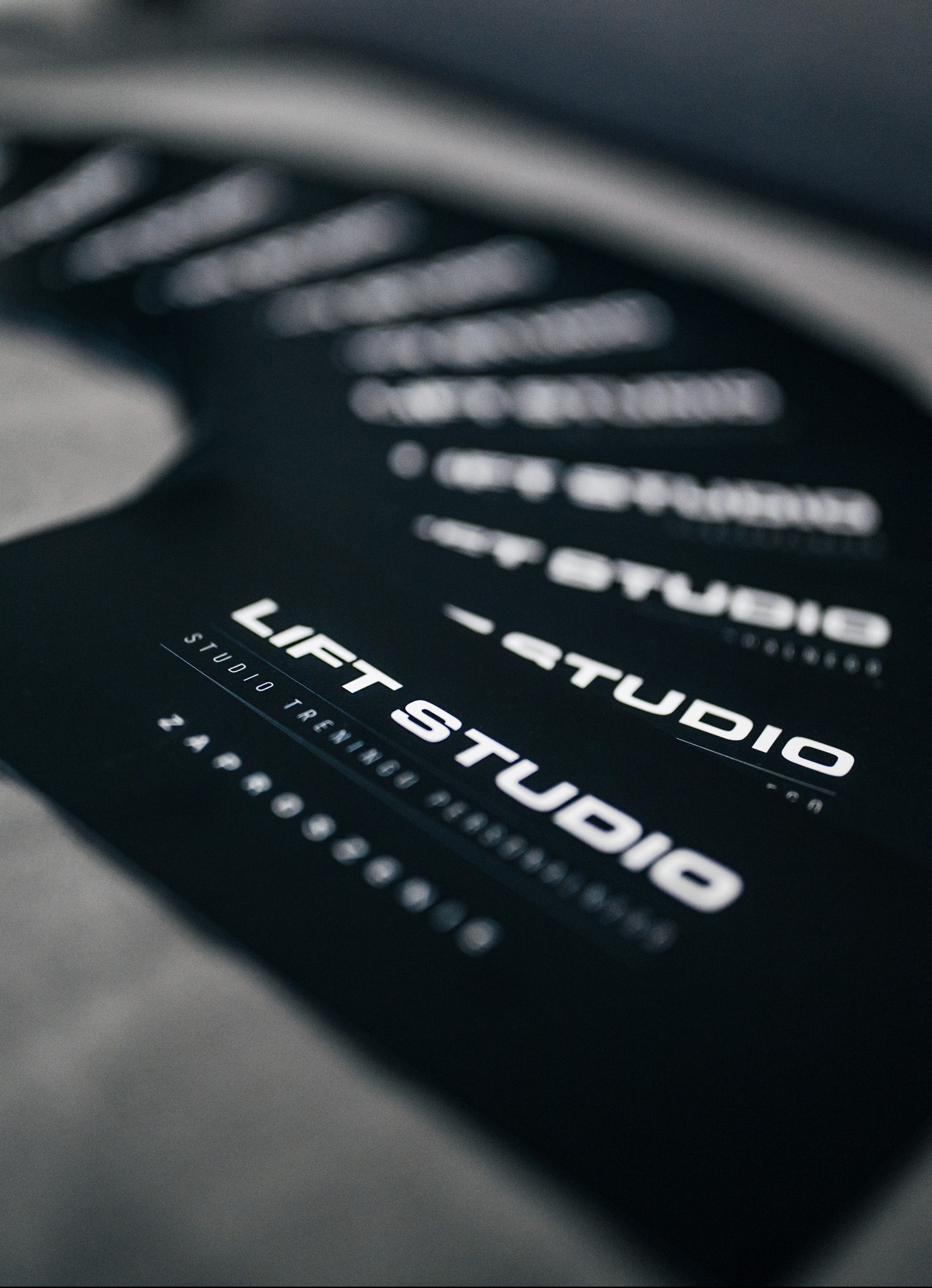 Stos wizytówek studia treningowego LIFT STUDIO, z białym tekstem na czarnym tle, ułożonych kaskadowo na szarym podłożu, z widocznym numerem telefonu.