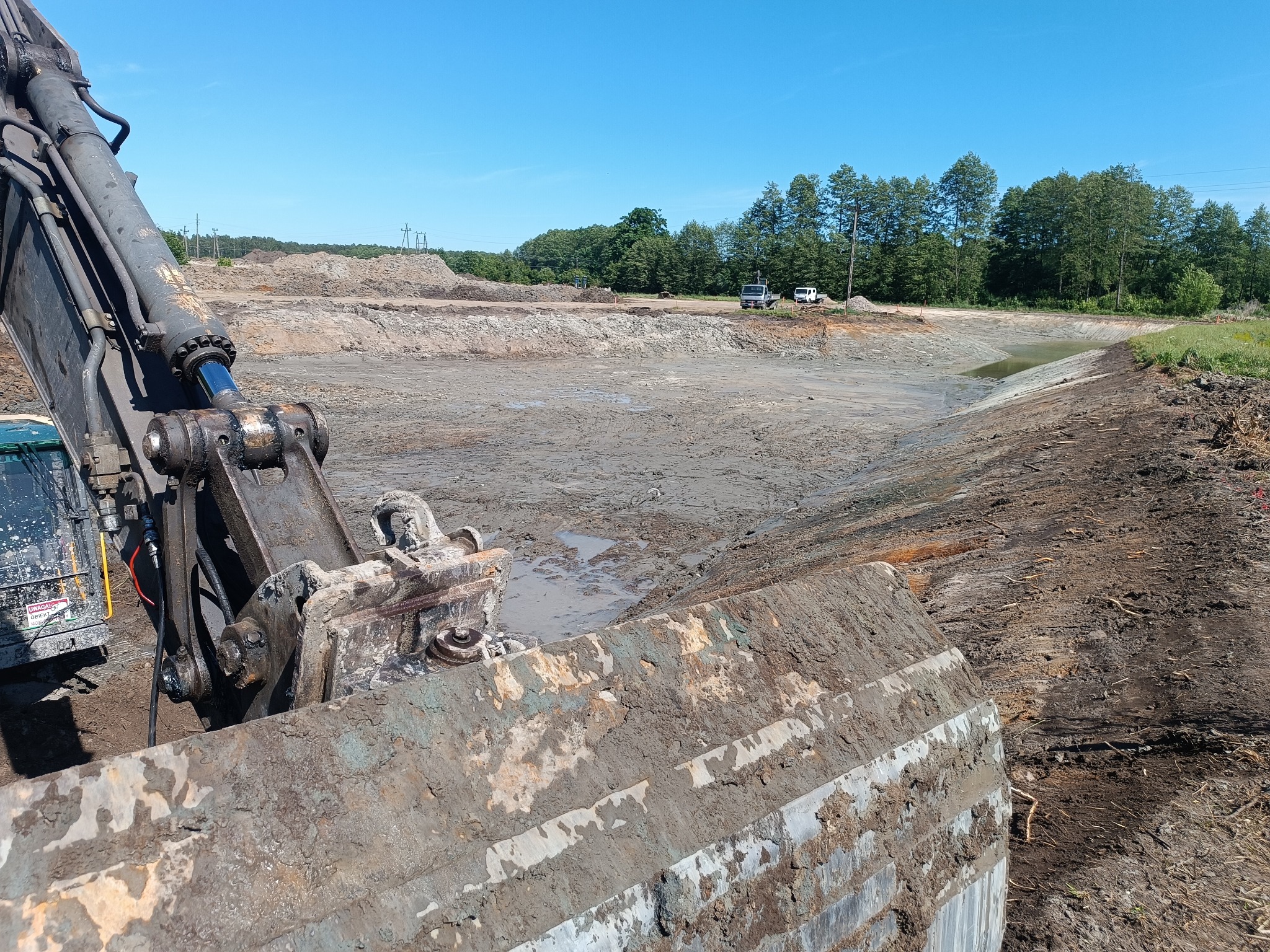 Głębinowy wykop pod staw, widok z kabiny koparki. Błękitne niebo, ziemia i woda. Ślady opon i gąsienic. W tle zieleń drzew i dwa białe samochody.