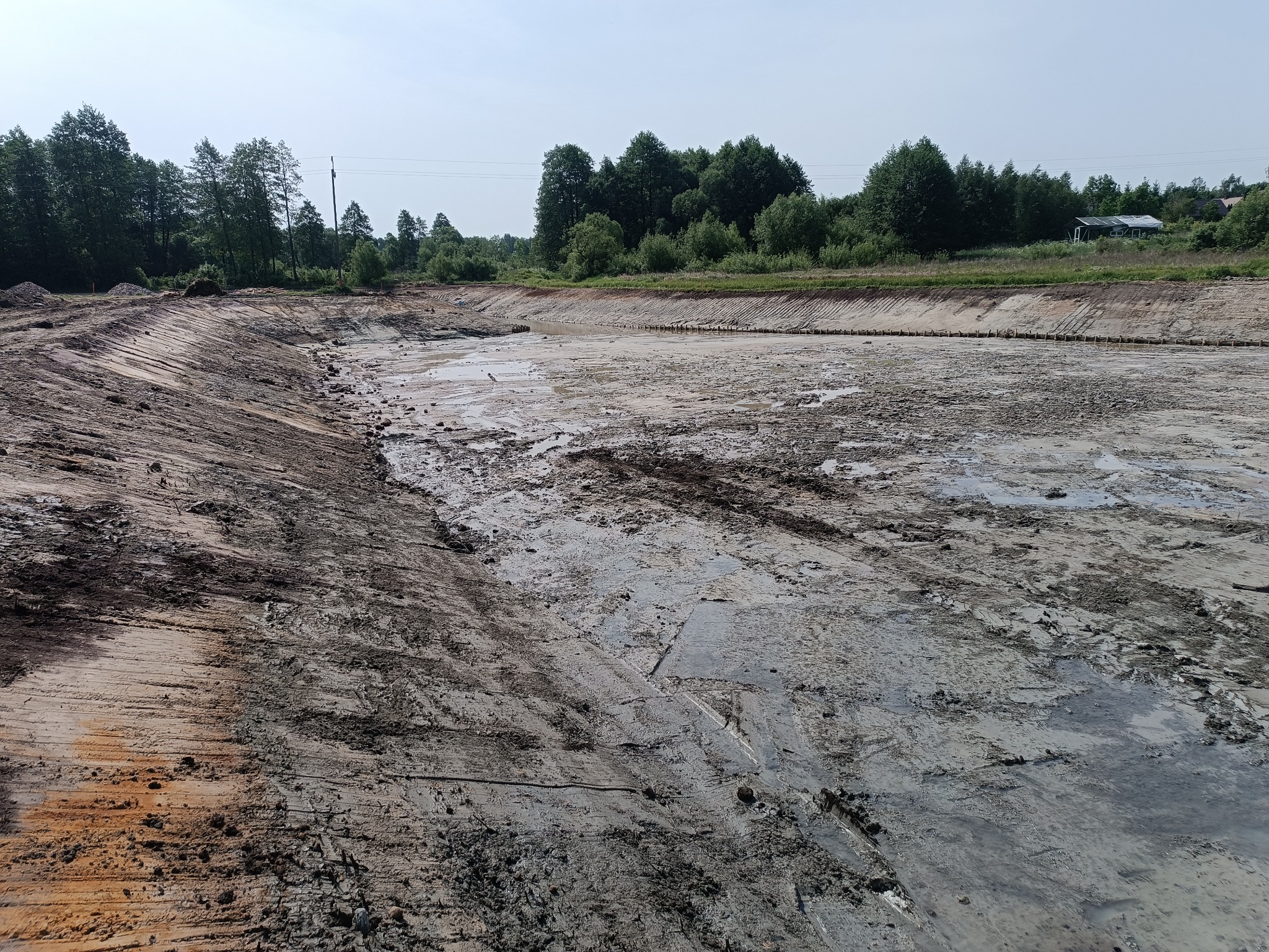 Wykopany, częściowo wypełniony wodą staw w trakcie prac ziemnych. Widoczne ślady po maszynach, ziemia i drzewa w tle. Brzegi stawu umocnione drewnianymi palikami.