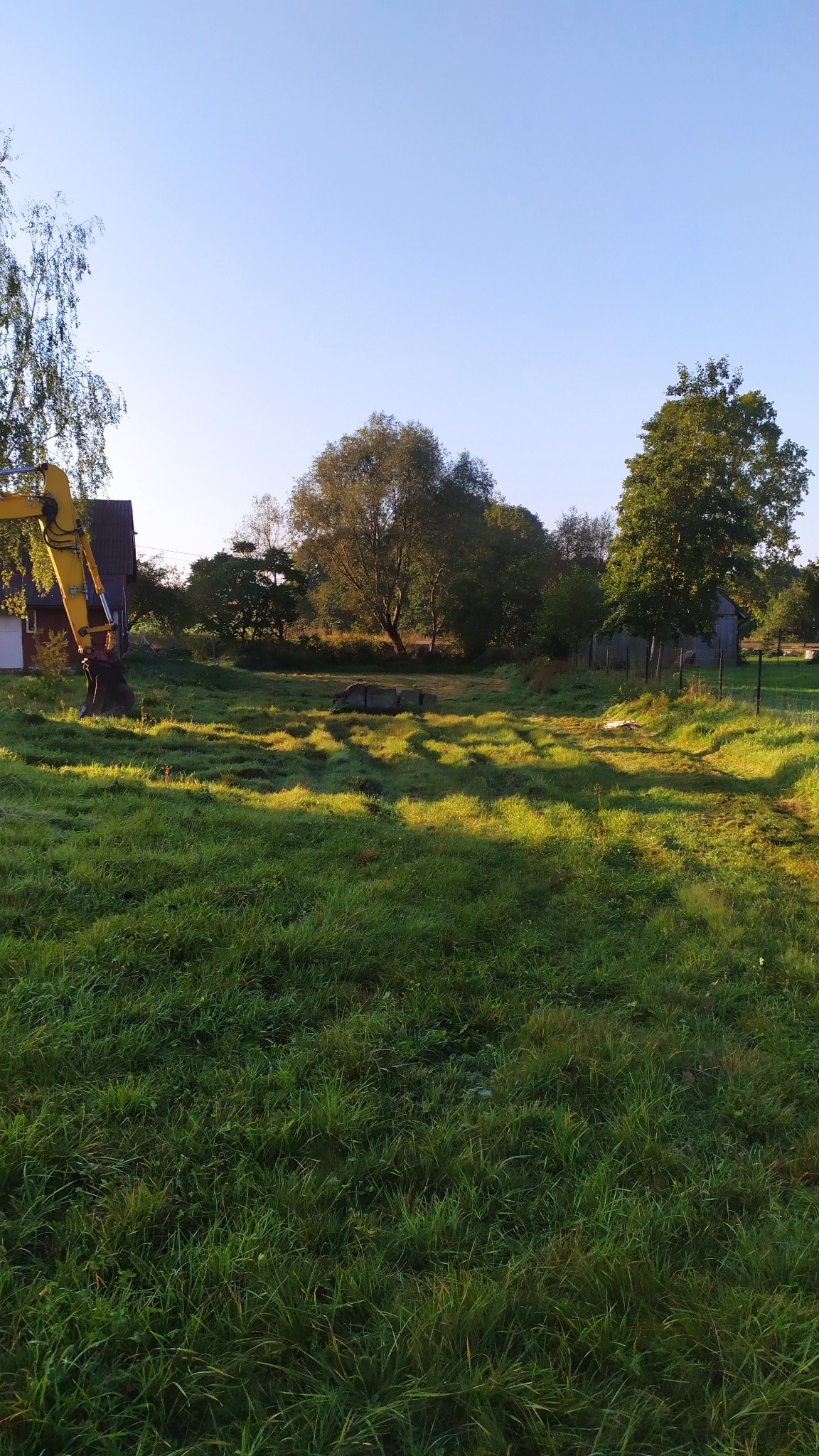 Żółta koparka na zroszonej łące, w tle drzewa i budynki. Poranne słońce oświetla trawę, tworząc malowniczy krajobraz. Widoczne betonowe elementy.