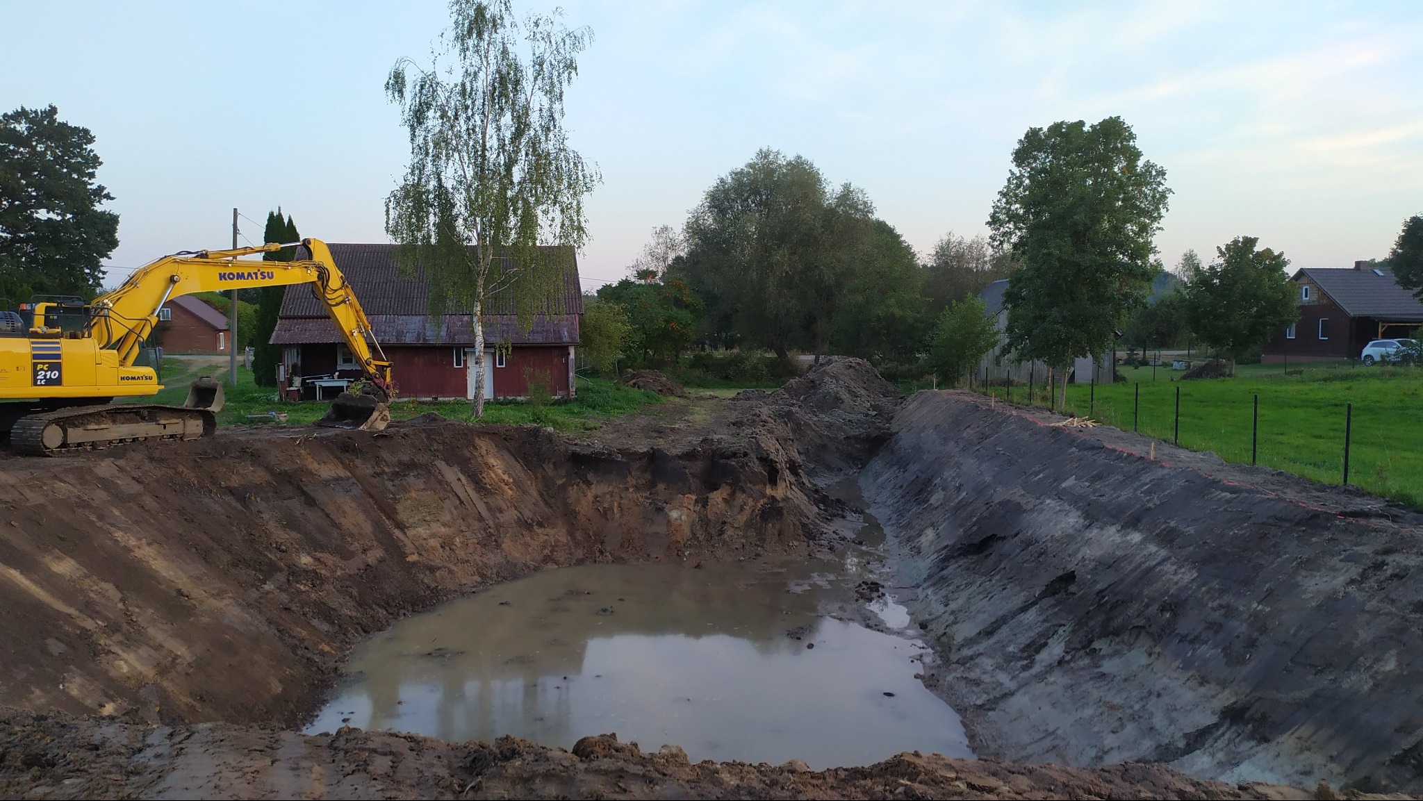 Żółta koparka Komatsu przy wykopanym stawie z wodą, obok czerwony budynek i drzewa. Wykopany staw w ziemi, widok z góry.