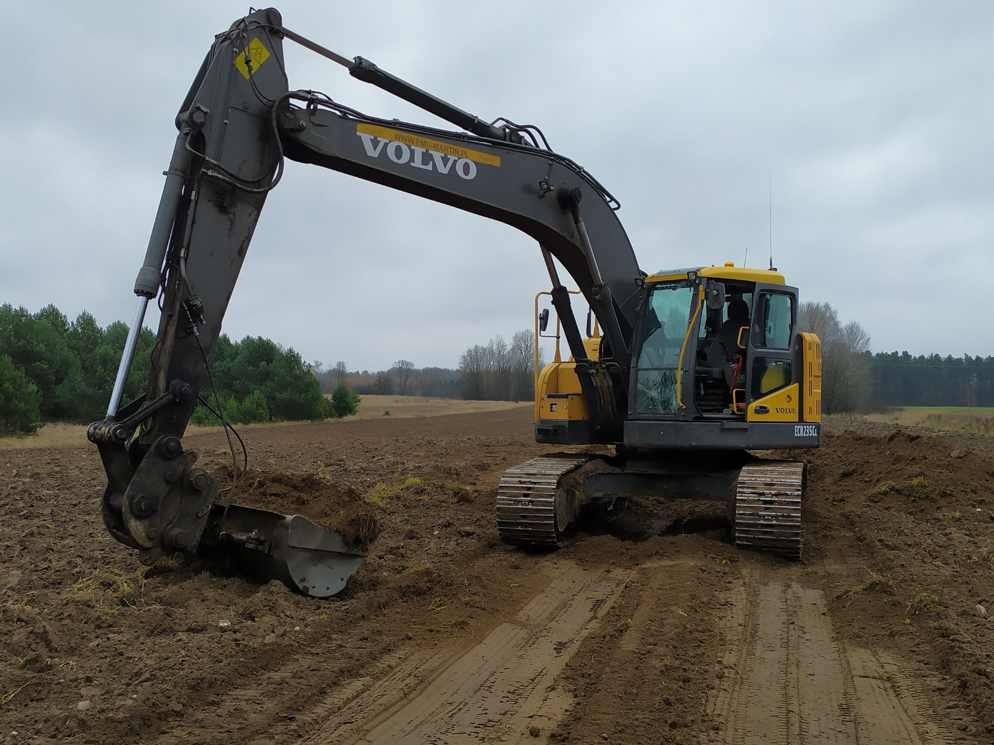 Żółto-szara koparka Volvo na polu, z widocznym łyżką zagłębiającą się w ziemię. W tle las i pochmurne niebo. Widoczne ślady opon.