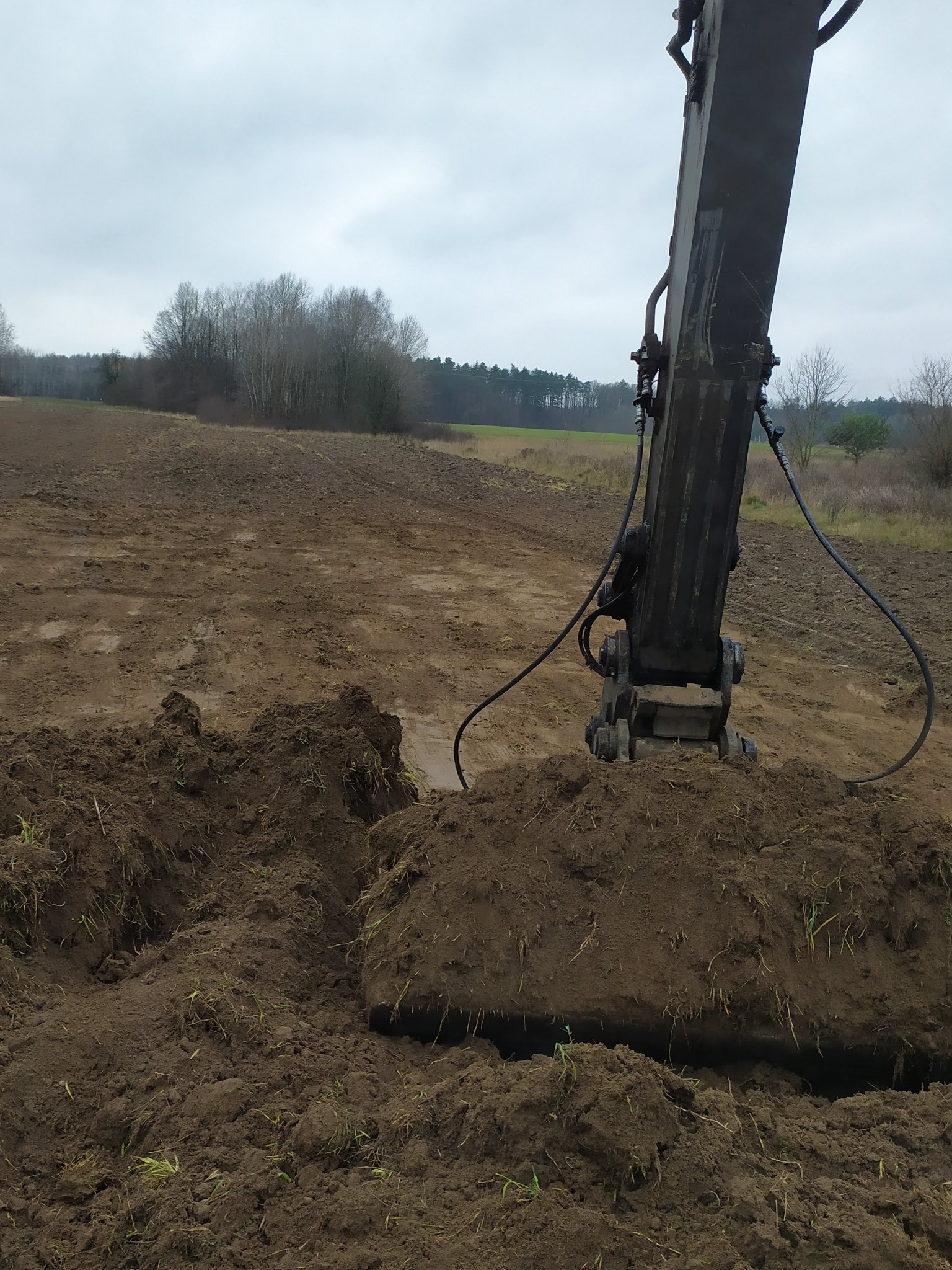 Koparka w trakcie prac ziemnych na polu, widoczny wykopany rów. Tło stanowią drzewa i pochmurne niebo. Prace ziemne w trakcie realizacji.