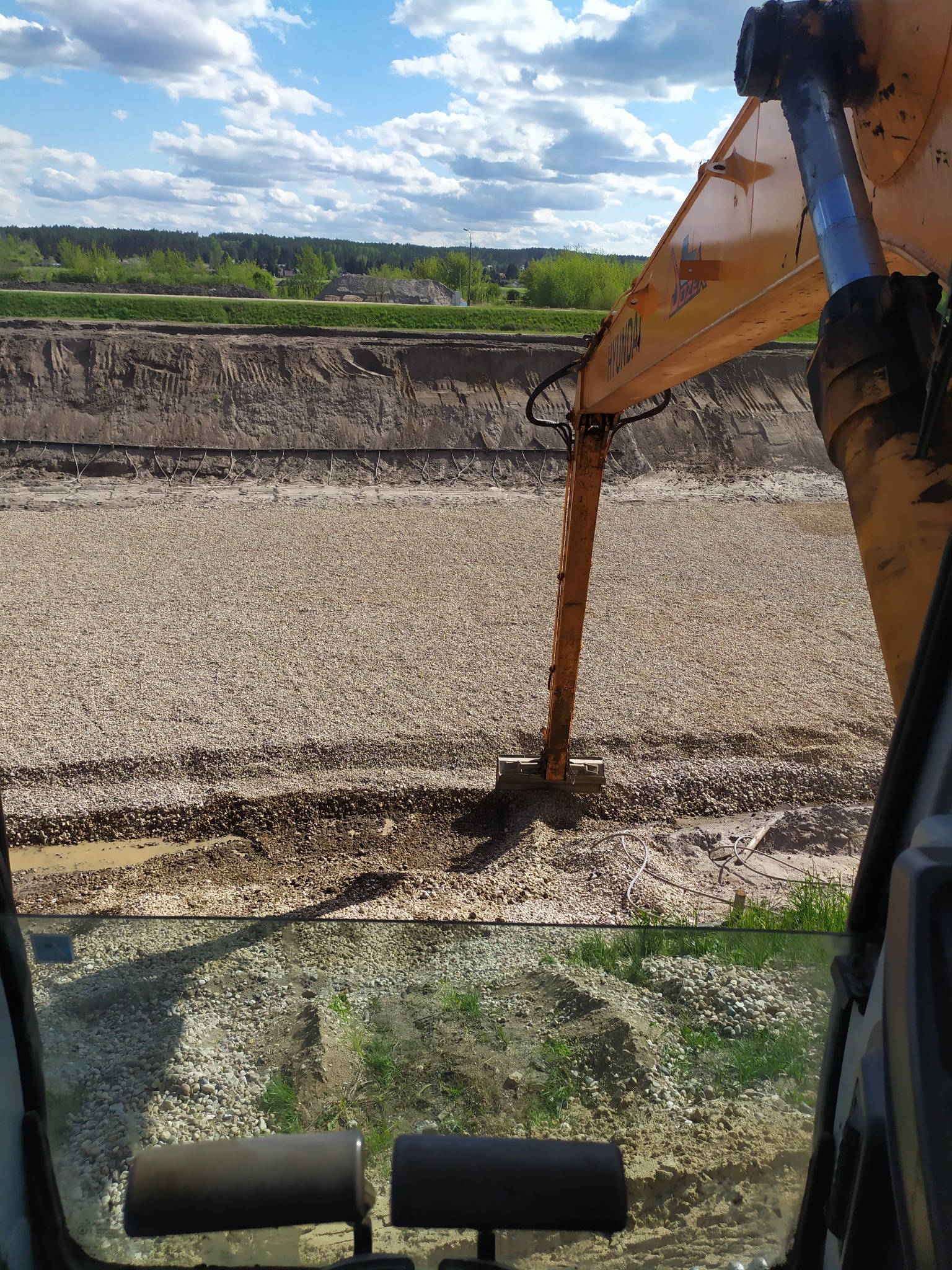 Widok z kabiny koparki Hyundai na wykopany i wysypany żwirem staw, w tle zielony krajobraz. Prace ziemne w toku, widoczne ślady pracy maszyny.
