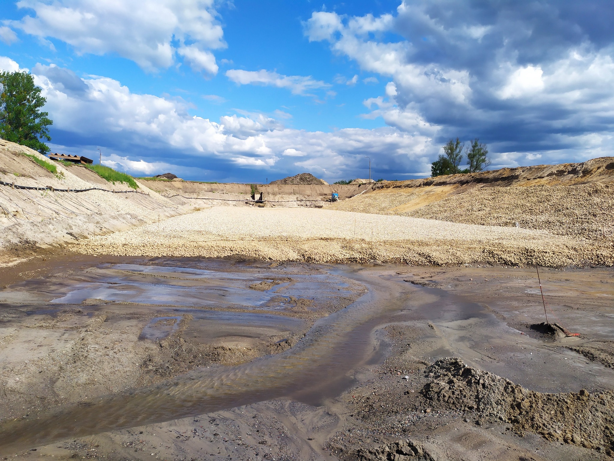 Szeroki kadr na wykopany staw z kamienistym brzegiem i wodą na dnie. Błękitne niebo z chmurami w tle. Widoczne ślady prac ziemnych i ukształtowanie terenu.
