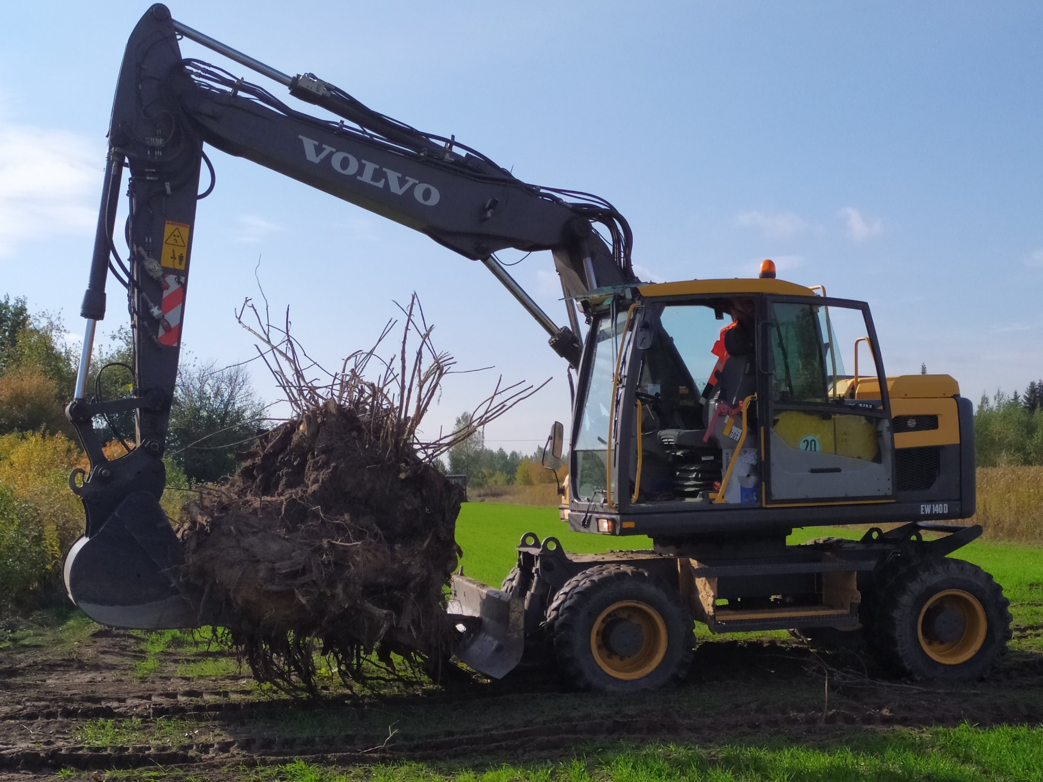 Żółto-czarna koparka Volvo EW140D z wyciągniętym pniem drzewa na tle zielonego pola i błękitnego nieba. Widoczne ślady opon na ziemi.
