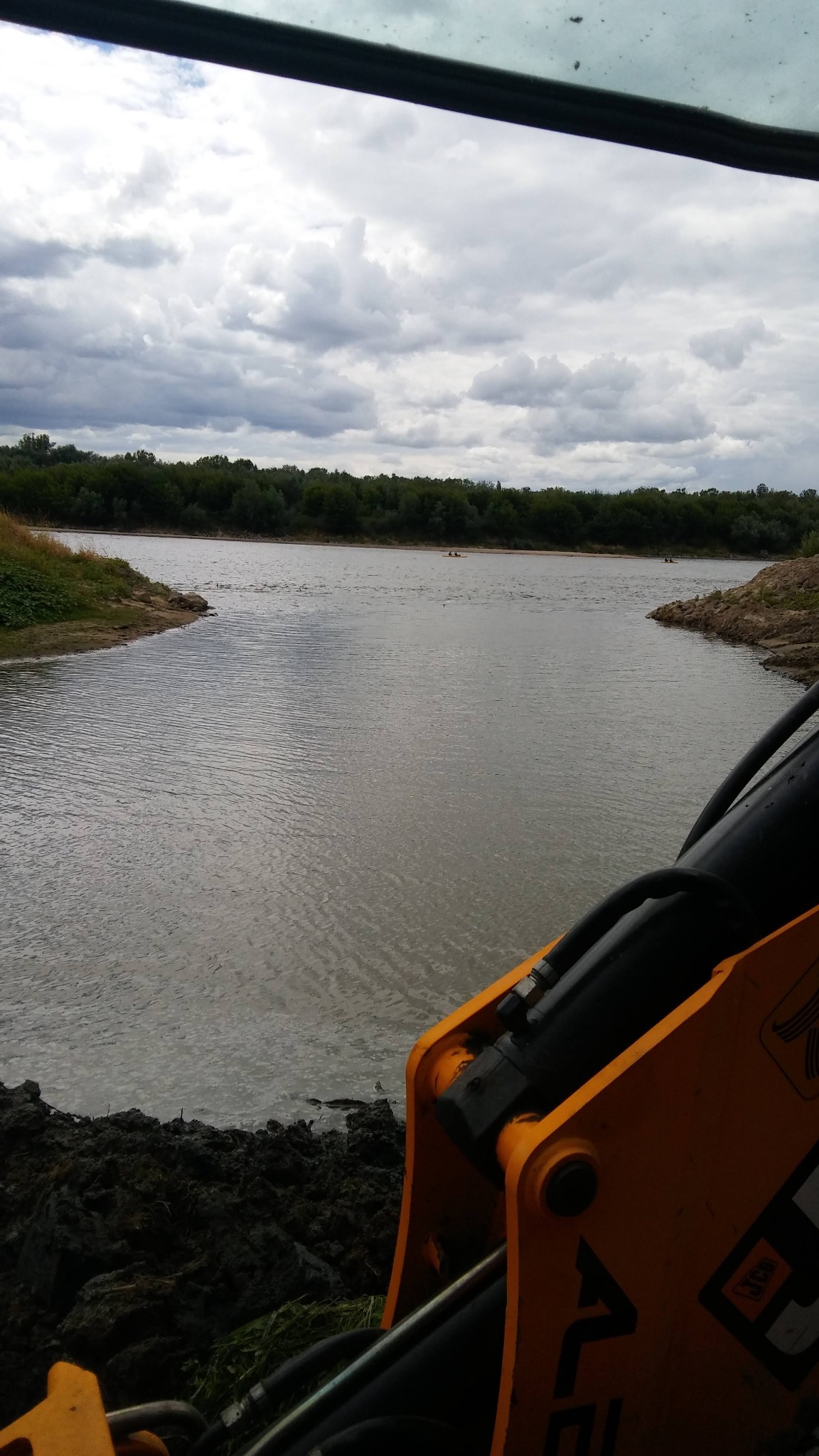 Widok z kabiny koparki na staw w trakcie prac ziemnych. W tle widoczne drzewa i pochmurne niebo. Żółte ramię koparki częściowo zasłania kadr.