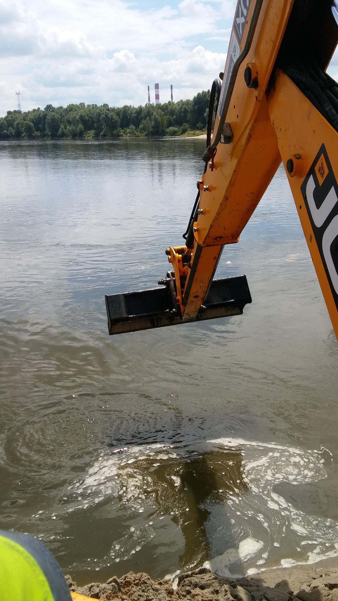 Żółta koparka JCB z czarną łyżką zanurzona w wodzie rzeki, w tle drzewa i kominy elektrowni. Prace ziemne w korycie rzeki, widoczne zmącenie wody i piana.