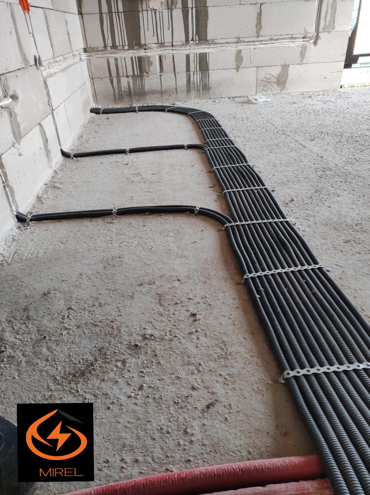 Równolegle ułożone rury instalacji elektrycznej w korycie, przymocowane do ściany w surowym stanie. Widoczne mocowania i fragment podłoża. Przewody w peszlu.