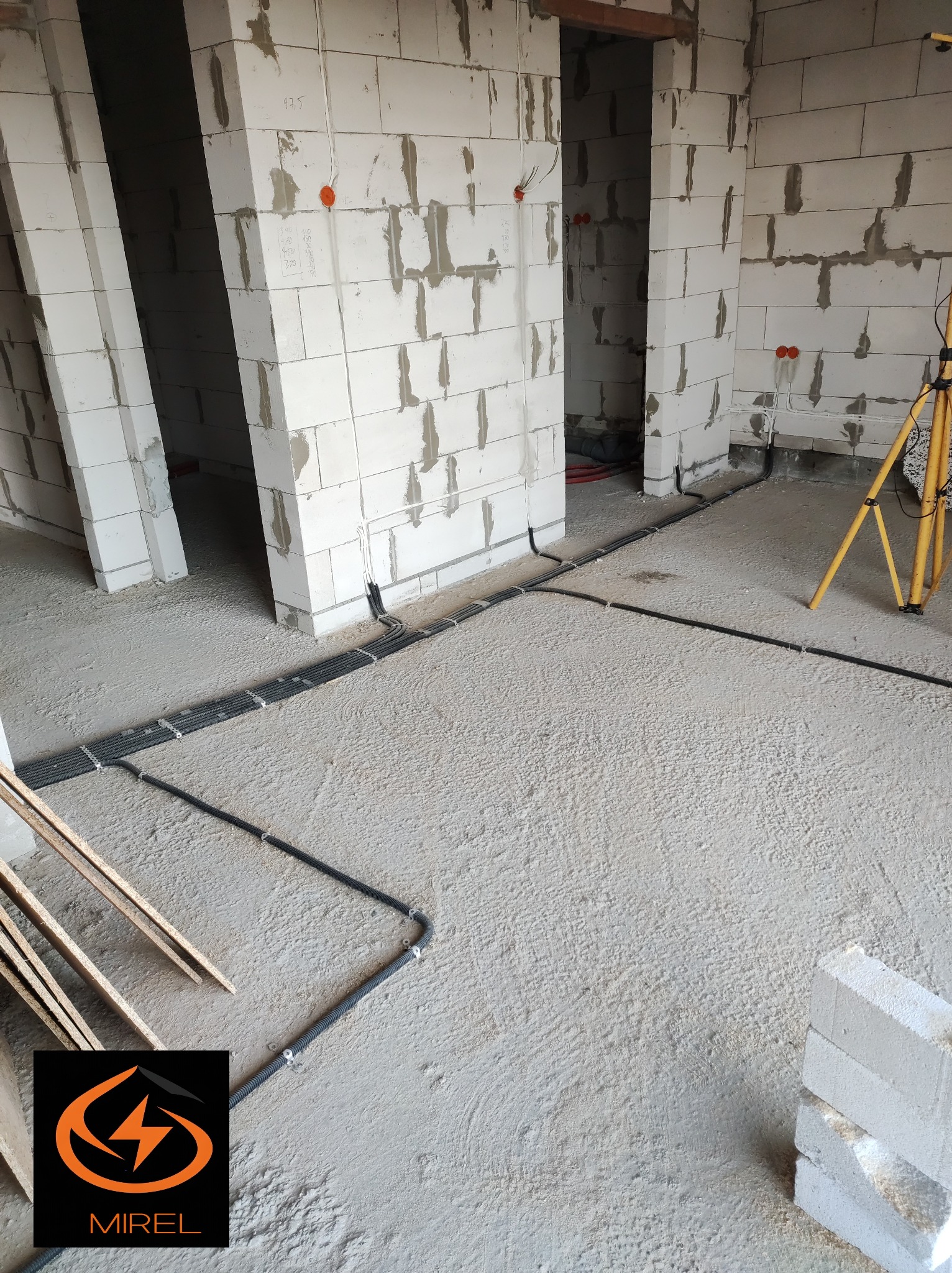 Surowy stan: kable elektryczne w peszlach ułożone na szlichtach, ściany z betonu komórkowego przed tynkowaniem. Widoczna lampa budowlana i logo firmy.