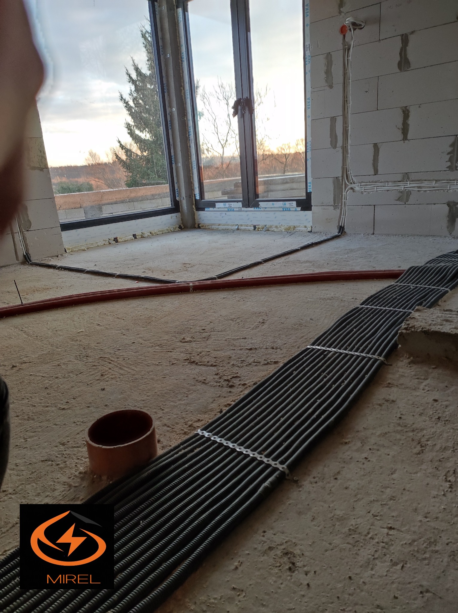 Wiązka kabli w peszlu ułożona na podłodze surowego wnętrza z oknem. Kable prowadzone wzdłuż ściany. Widoczne przewody instalacji elektrycznej w ścianie.