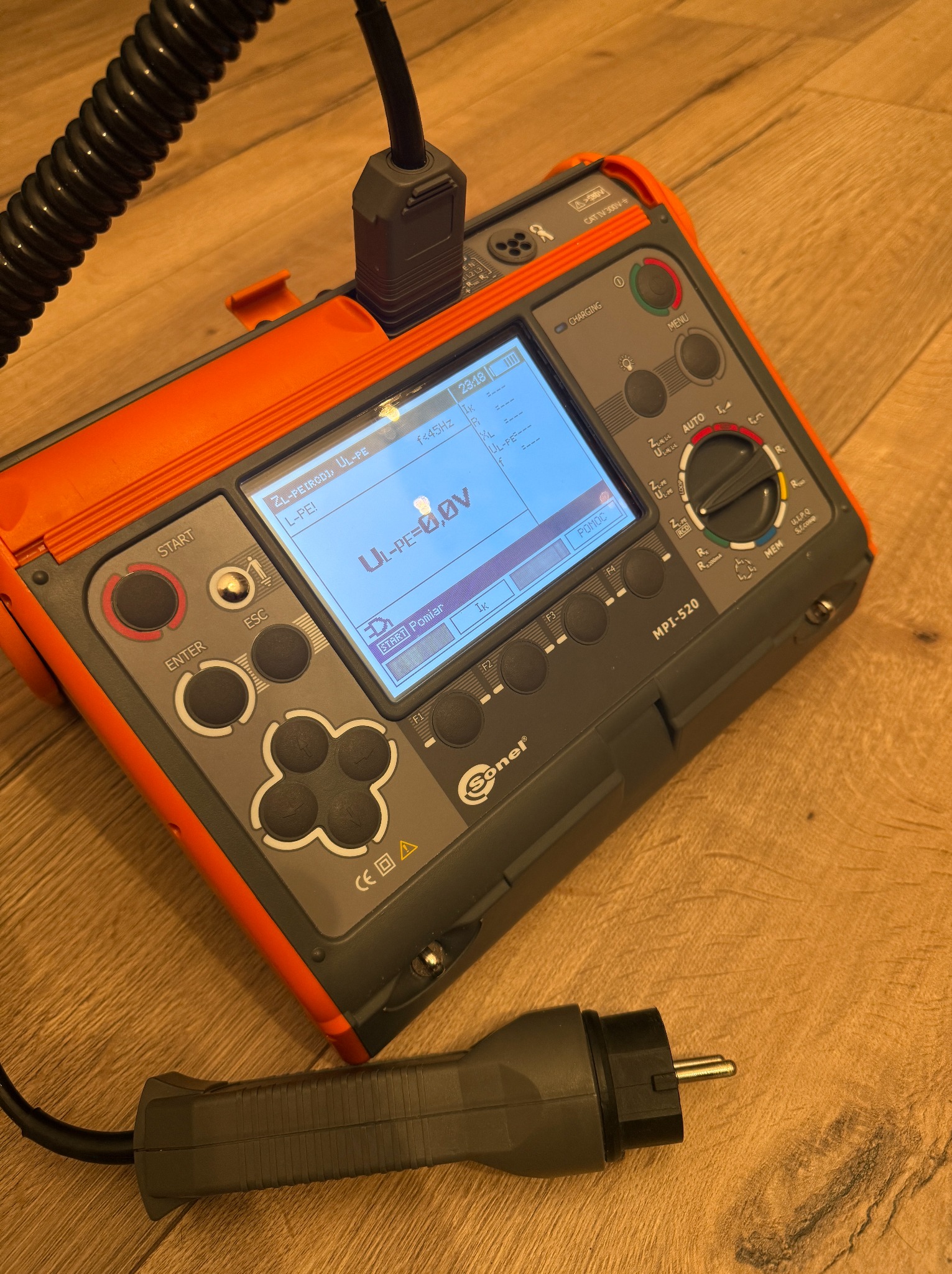 Miernik MPI-520 Sonel z podłączonym przewodem i wtyczką, wyświetlający parametry pomiarowe na ekranie LCD. Urządzenie leży na drewnianej powierzchni.