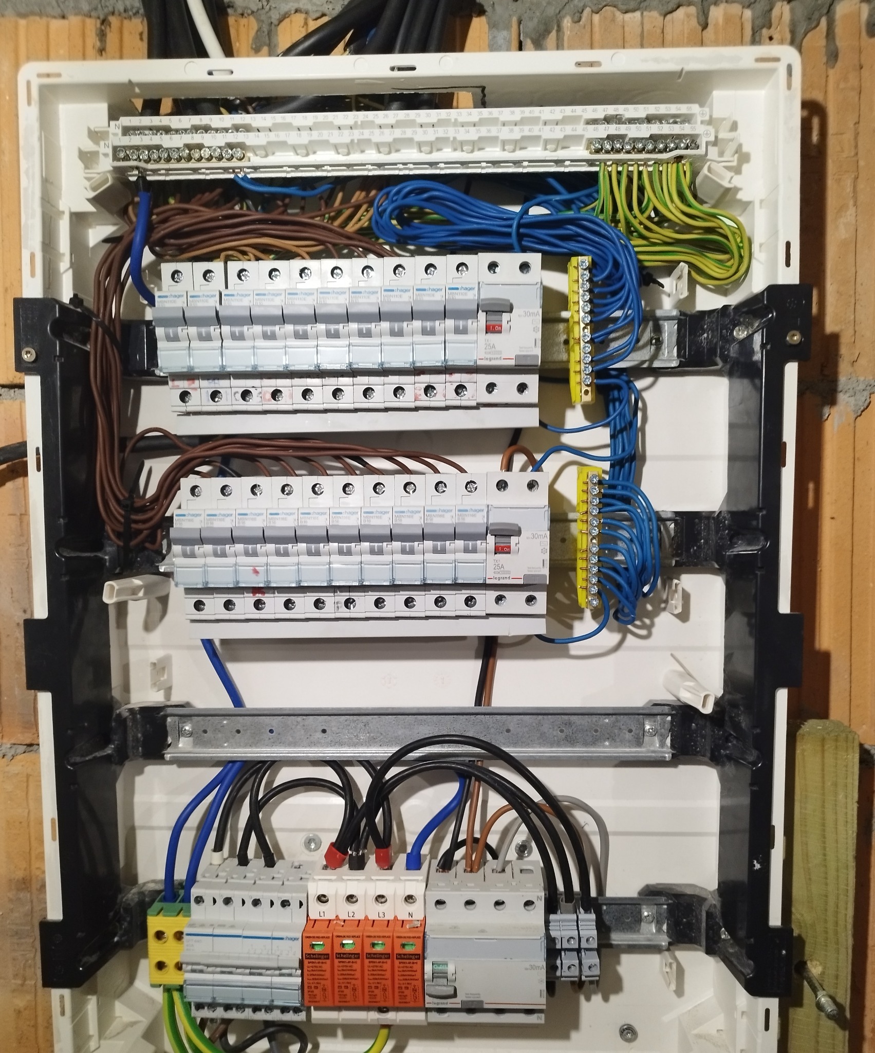 Rozdzielnica elektryczna z zainstalowanymi bezpiecznikami, przewodami w różnych kolorach i zabezpieczeniami przeciwprzepięciowymi, zamontowana na ceglanej ścianie.