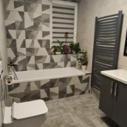 Wykończona łazienka z geometrycznymi płytkami w odcieniach szarości, białą wanną i toaletą, grzejnikiem drabinkowym oraz szafką z umywalką; na parapecie okna widoczne rośliny doniczkowe.