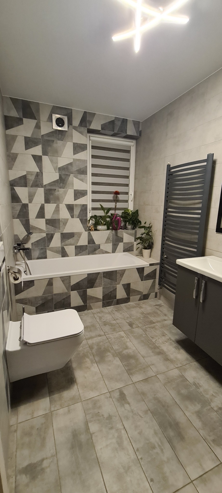 Wykończona łazienka z geometrycznymi płytkami w odcieniach szarości, białą wanną i toaletą, grzejnikiem drabinkowym oraz szafką z umywalką; na parapecie okna widoczne rośliny doniczkowe.