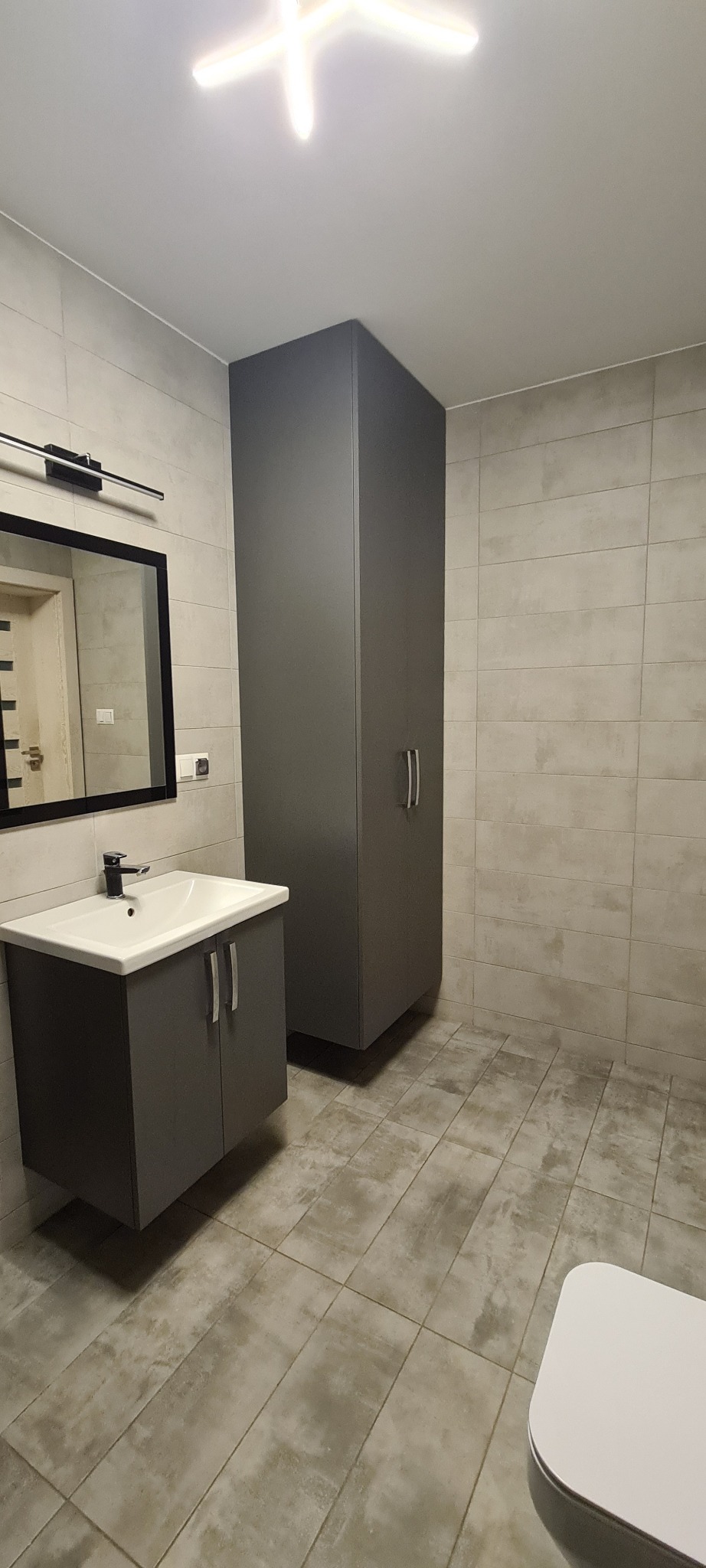 Nowoczesna łazienka w szarościach z wiszącą szafką umywalkową, wysoką szafką i dużym lustrem w czarnej ramie, oświetlona plafonem.