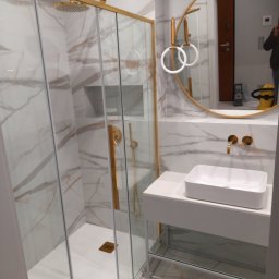 Kadek Usługi Remontowo-Wykończeniowe - Nowoczesna łazienka z kabiną prysznicową ze złotymi ramami, białą umywalką nablatową, okrągłym lustrem i marmurowymi płytkami na ścianach i podłodze.