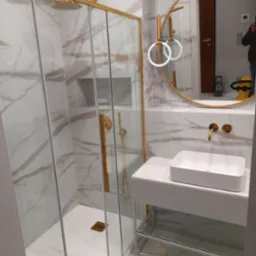 Nowoczesna łazienka z kabiną prysznicową ze złotymi ramami, białą umywalką nablatową, okrągłym lustrem i marmurowymi płytkami na ścianach i podłodze.