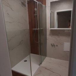 Kadek Usługi Remontowo-Wykończeniowe - Nowoczesna łazienka z kabiną prysznicową typu walk-in, wykończona marmurowymi płytkami w odcieniach beżu i brązu, z akcentem drewnianej ściany za kabiną i drewnianą podłogą w części wejściowej.