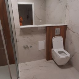 Kadek Usługi Remontowo-Wykończeniowe - Nowoczesna łazienka z kabiną prysznicową, toaletą podwieszaną i lustrem, wykończona płytkami imitującymi marmur.